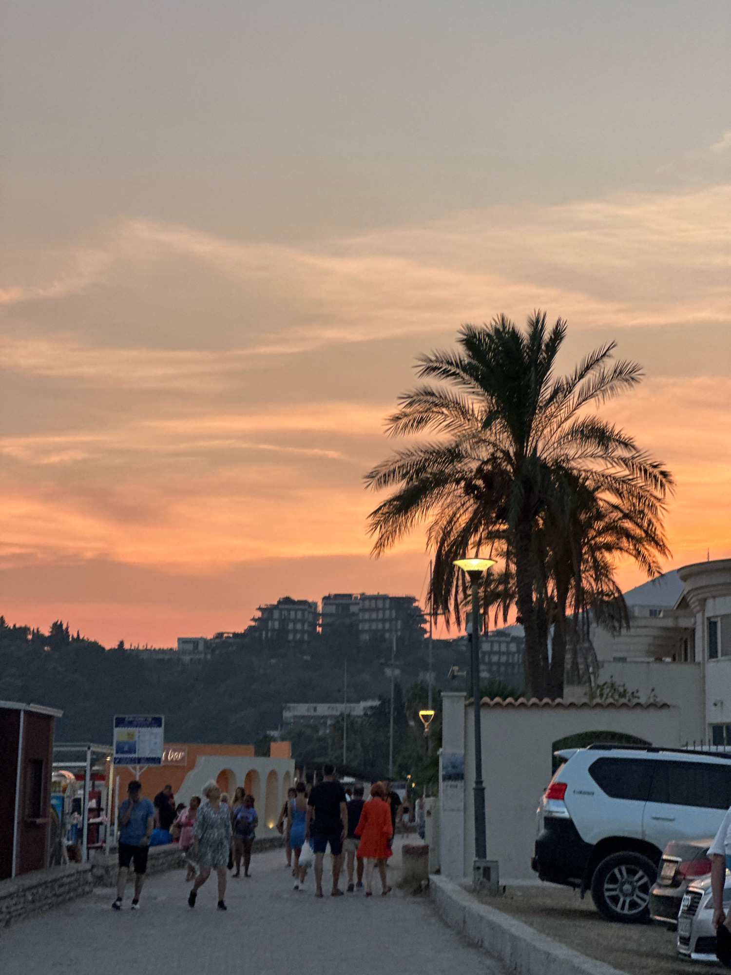 The image depicts a street scene in Boreti, Montenegro, at dusk. The sky is a gradient of orange and pale blue, with scattered clouds. A large palm tree dominates the center-right of the frame, its fronds silhouetted against the sky. In the background, several buildings are visible on a hillside, and some modern apartment buildings are also present.
In the foreground, a paved walkway runs horizontally across the image. Several people are walking along the path. On the left, a red-roofed structure with an open front is visible. On the right side of the walkway, a white SUV is parked, its rear end facing the camera. Further along the walkway, a street lamp is illuminated, casting a soft glow.
The people on the walkway are dressed in casual summer clothing, suggesting a warm evening. Some are walking alone, while others appear to be in groups. Their actions are varied, with some looking at their phones, others engaged in conversation. The overall atmosphere is relaxed and serene, characteristic of a seaside town at the end of the day.