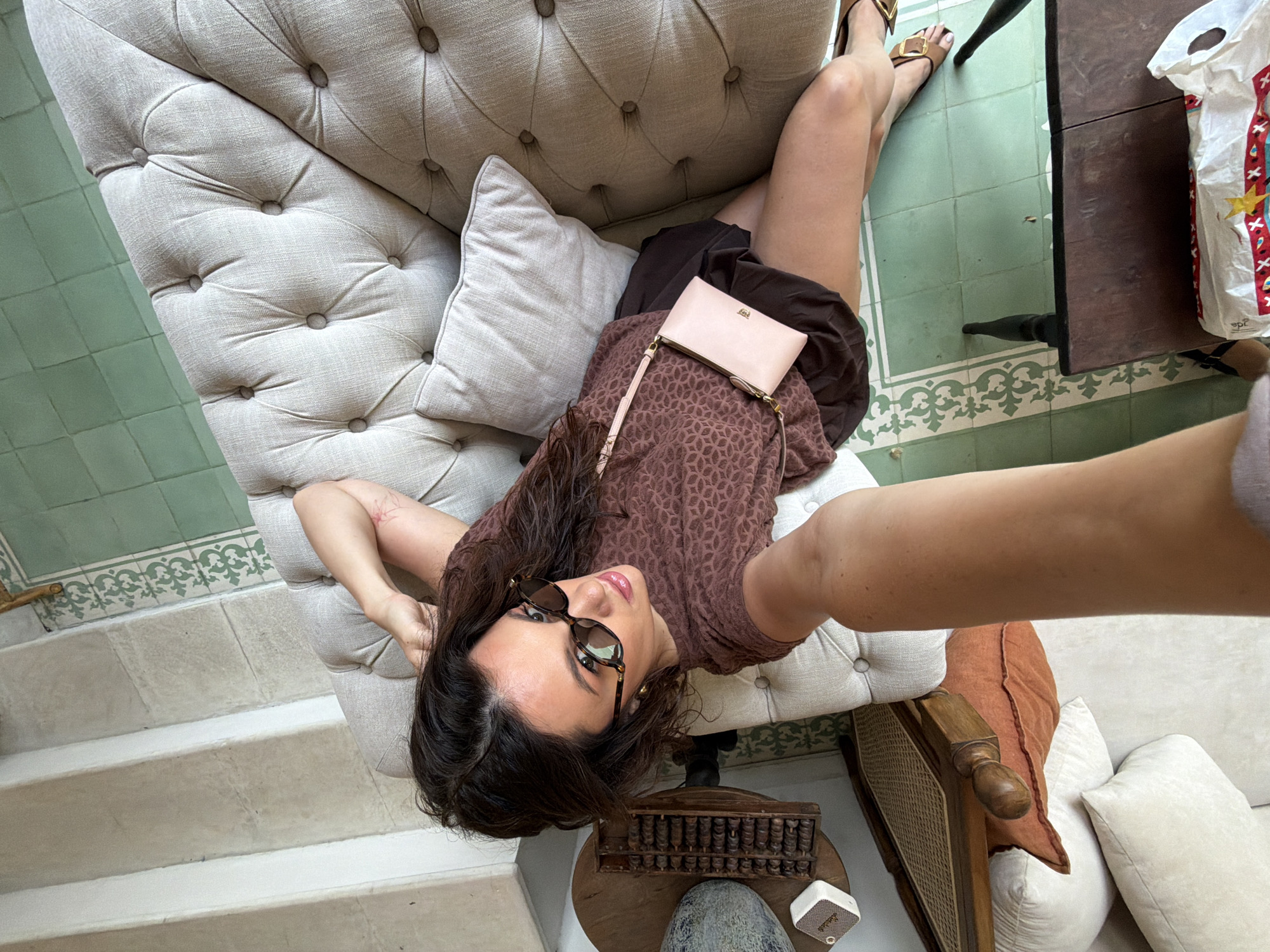 A medium-dark skinned woman with long, dark, wavy hair and wearing sunglasses is taking a selfie while sitting on a tufted light-colored sofa. She is wearing a brown patterned short-sleeved shirt and dark brown shorts. A light pink purse with a thin strap is slung across her body. Her right arm is resting on the back of the sofa. To her left, there is a dark wooden table with a white shopping bag on it. The floor is tiled with green squares and white decorative trim. In the background, there are stairs leading upwards and other furniture, including a wooden chair with woven material and cushions. The overall environment appears to be an indoor space with natural light.