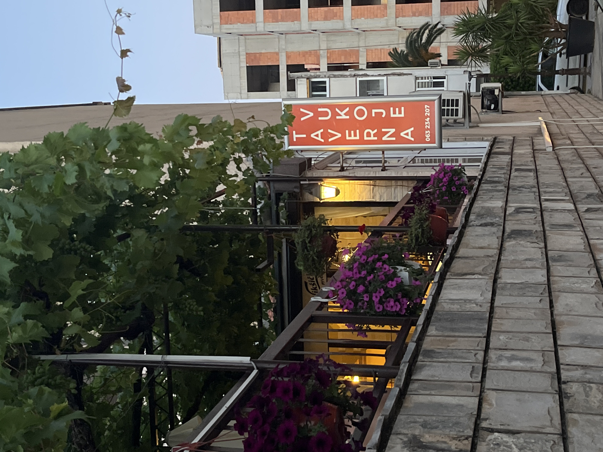 The image depicts an outdoor scene in Budva, Montenegro, featuring a restaurant facade with floral decorations. A vertical, orange sign with white lettering reads "Taverna Vukoje" and includes a phone number, "063 234 207". The sign is illuminated by a nearby lamp. Several potted plants with purple flowers hang from a railing, adding to the inviting atmosphere. In the background, buildings under construction are visible, with concrete and brick elements. Green foliage from a tree or vine drapes over the scene. The ground in the foreground is paved with stone. A white car is parked on the street to the right, with a palm tree and other greenery in planters. The sky is a pale blue, suggesting either early morning or late evening.
