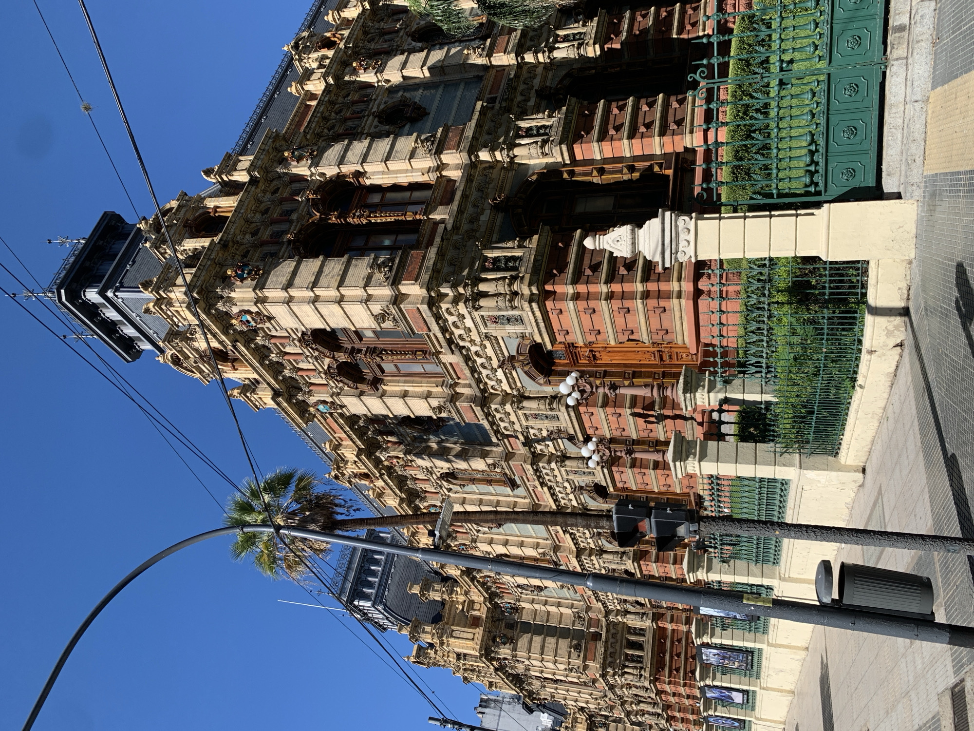 This media shows a grand, highly ornate building on a sunny street in what appears to be an urban setting. The multi-story building features elaborate architecture, combining light-colored stone or stucco with reddish brickwork and dark wood elements. Its facade is richly detailed with intricate carvings, pilasters, cornices, decorative balconies, and numerous arched and rectangular windows. The upper levels include dormer windows and a dark, possibly slate, roof with further decorative accents.
At street level, a dark green, ornate wrought-iron fence and gate delineate the property, with neatly trimmed hedges visible behind it. A light-colored stone pillar marks the entrance. The sidewalk is paved with distinctive patterned tiles, predominantly gray with yellow accents, characteristic of Buenos Aires.
In the foreground, a tall, dark street light pole stands prominently, equipped with a traffic light and what appear to be small, unreadable posters or advertisements attached. Overhead utility wires crisscross the clear, bright blue sky. A large palm tree with a thick trunk and green fronds adds to the streetscape, casting some shadow.
The scene is bathed in bright, direct sunlight, creating strong contrasts and shadows that emphasize the architectural textures and details. The time of day is clearly daytime, under a cloudless sky, suggesting fair weather. The street is quiet, with no people or vehicles visible, depicting a static moment of this architecturally significant location in Buenos Aires, Argentina.