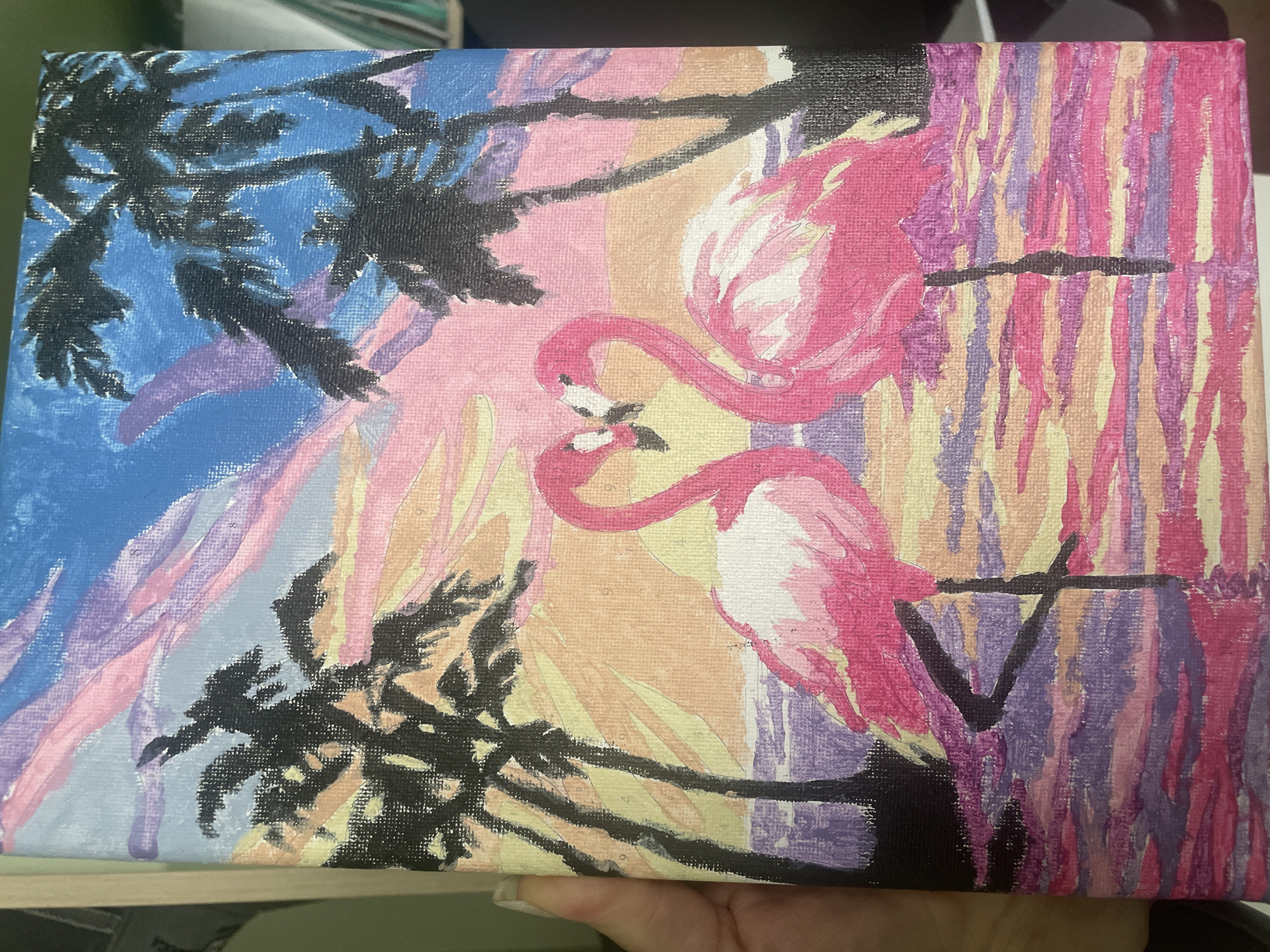 The image is a close-up shot of a paint-by-numbers style painting on canvas. The painting depicts two pink flamingos with their necks intertwined, set against a stylized sunset or sky. Palm tree silhouettes frame the scene on the left and right. The color palette includes shades of blue, pink, purple, yellow, and orange. The painting has a somewhat unfinished or amateurish quality, with visible brush strokes and numbered sections of the canvas that need to be painted. The background consists of vertical stripes of varying colors. The canvas is held by a hand. There is no evidence to suggest that the image was taken in Saratov, Russia.