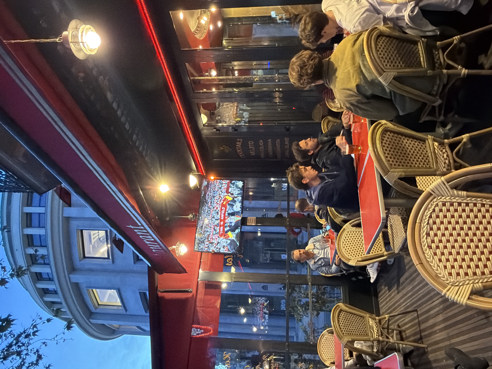 The image depicts a scene at a restaurant in Paris, France, at dusk. Several people are seated at tables along the exterior of the establishment. Some appear to be watching a television screen visible through the restaurant's glass exterior. Tables and chairs are arranged in a typical outdoor cafe setting. A red awning covers the seating area. The menu, visible on a glass door, includes cocktails such as mojitos, piña coladas, and margaritas. The building across the street has a distinctive curved facade. The scene suggests a casual dining or social gathering with patrons engaging in conversation and watching a sporting event.