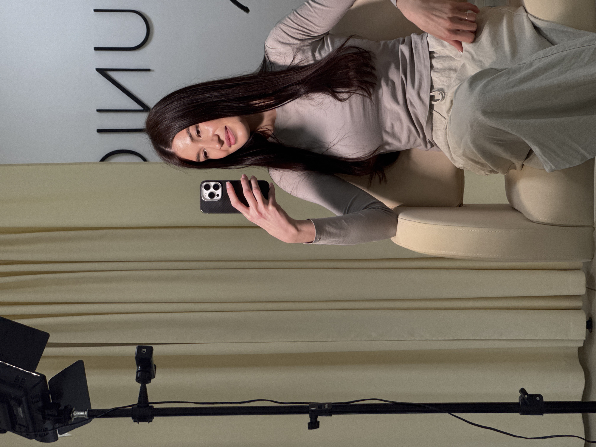 A woman with long, dark hair is sitting on a light-beige couch and taking a selfie with a black smartphone. She is wearing a light-gray long-sleeved shirt and light-beige pants. Behind her, a white wall has the word "CLINIC" written in black, sans-serif, stylized letters. Below the wall, light-beige curtains are hung. In the lower portion of the image, a professional lighting setup with a light stand and a black light is visible. The photo appears to have been taken indoors, likely in a studio or office setting, though there are no clues that identify Chaikovsky, Russia.