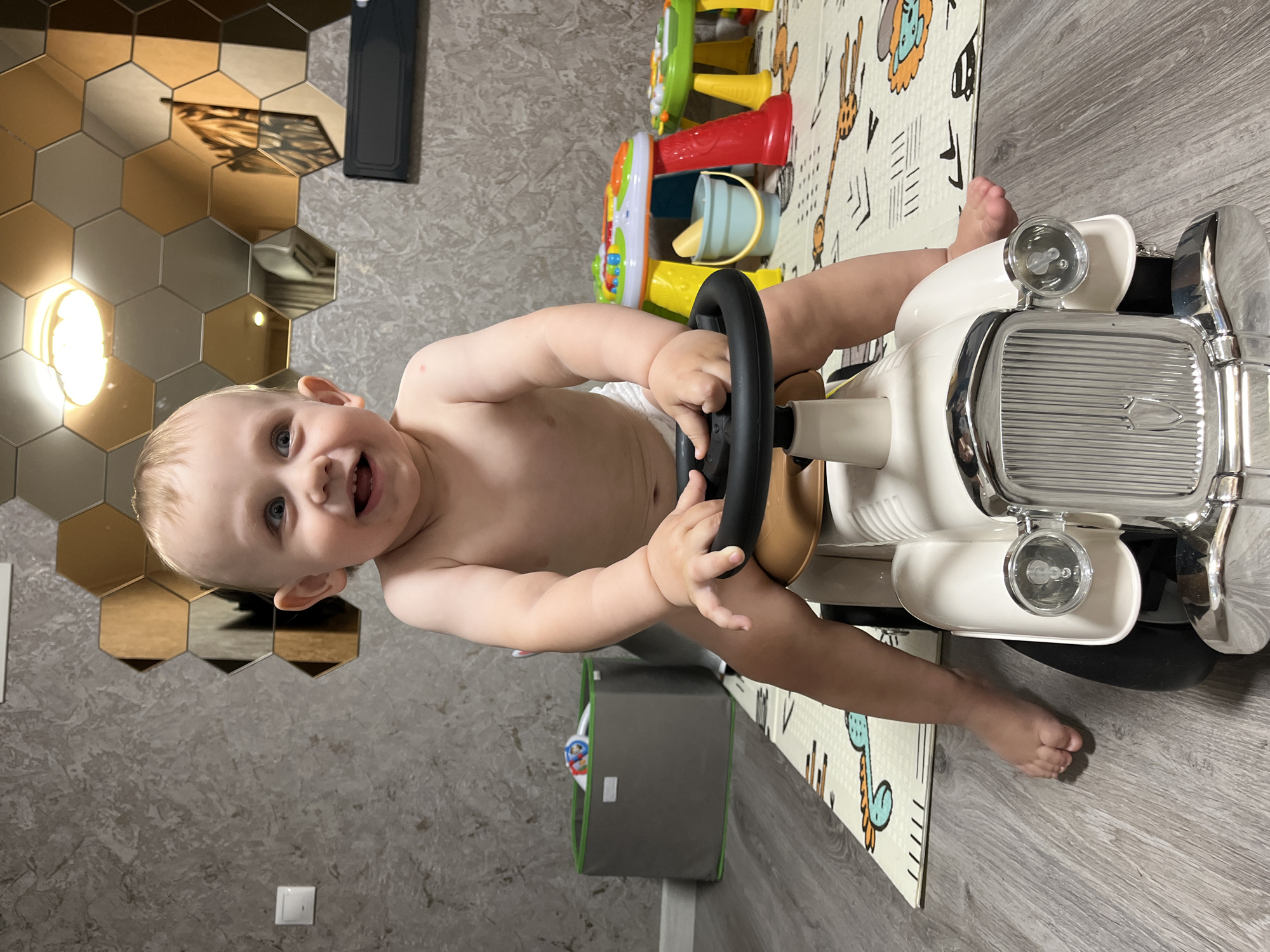 A baby is playing with a white toy car. The baby is smiling and sitting inside the car. The car has a steering wheel and a grill. The baby is wearing a diaper. There are toys in the background, including a toy kitchen. The scene is indoors. The floor is wooden. There are hexagonal mirrors on the wall. The baby looks happy and engaged. The lighting appears to be natural, suggesting it might be daytime. The overall setting is a playroom or a child's room.
