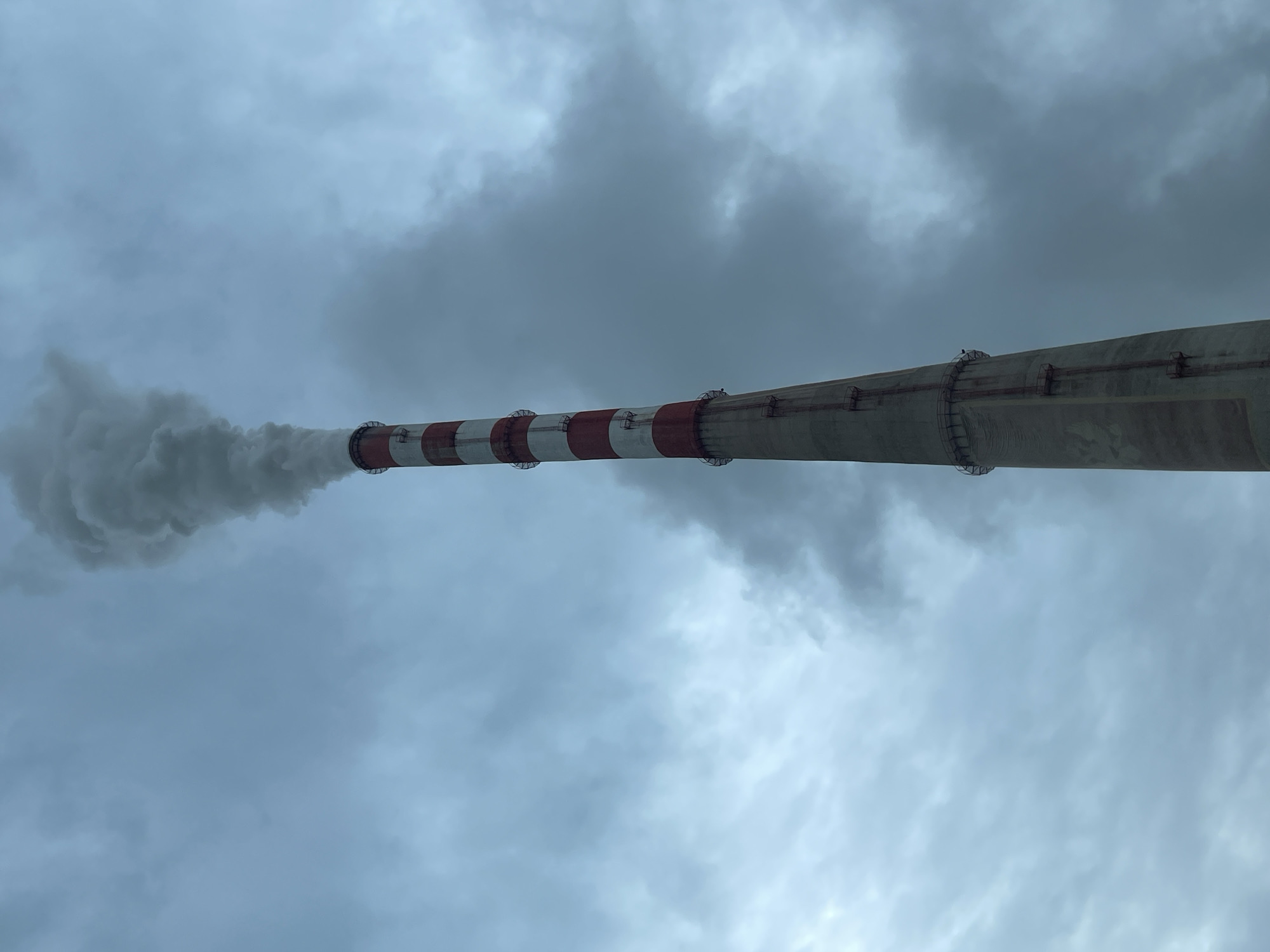 A horizontal shot of a tall, thin smokestack, striped red and white at the top, extending into a cloudy, overcast sky. Plumes of smoke billow out of the top of the stack. The smokestack is located in Kalušići, Montenegro.