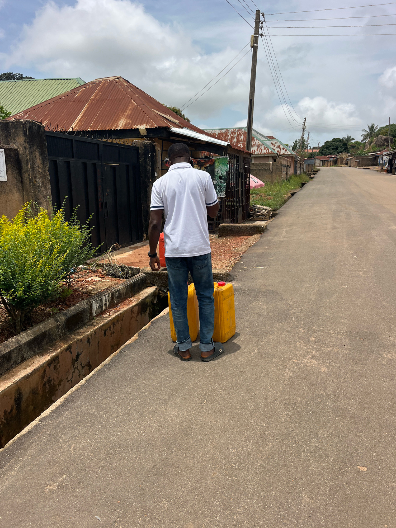A man, viewed from behind, walks down a paved street in Gura Top, Nigeria, carrying two large yellow plastic jerrycans. He wears a white polo shirt with blue trim and blue jeans, and dark sandals. The jerrycans have red caps. To the left of the man is a drainage ditch, and beyond that, a low concrete wall topped with a black metal gate and a rusty corrugated metal roof on a building. Lush green bushes are planted to the left of the gate. The street continues into the distance, lined with various buildings with corrugated metal roofs and some with cinder block walls. Electrical wires are strung across poles along the street. The sky is cloudy.