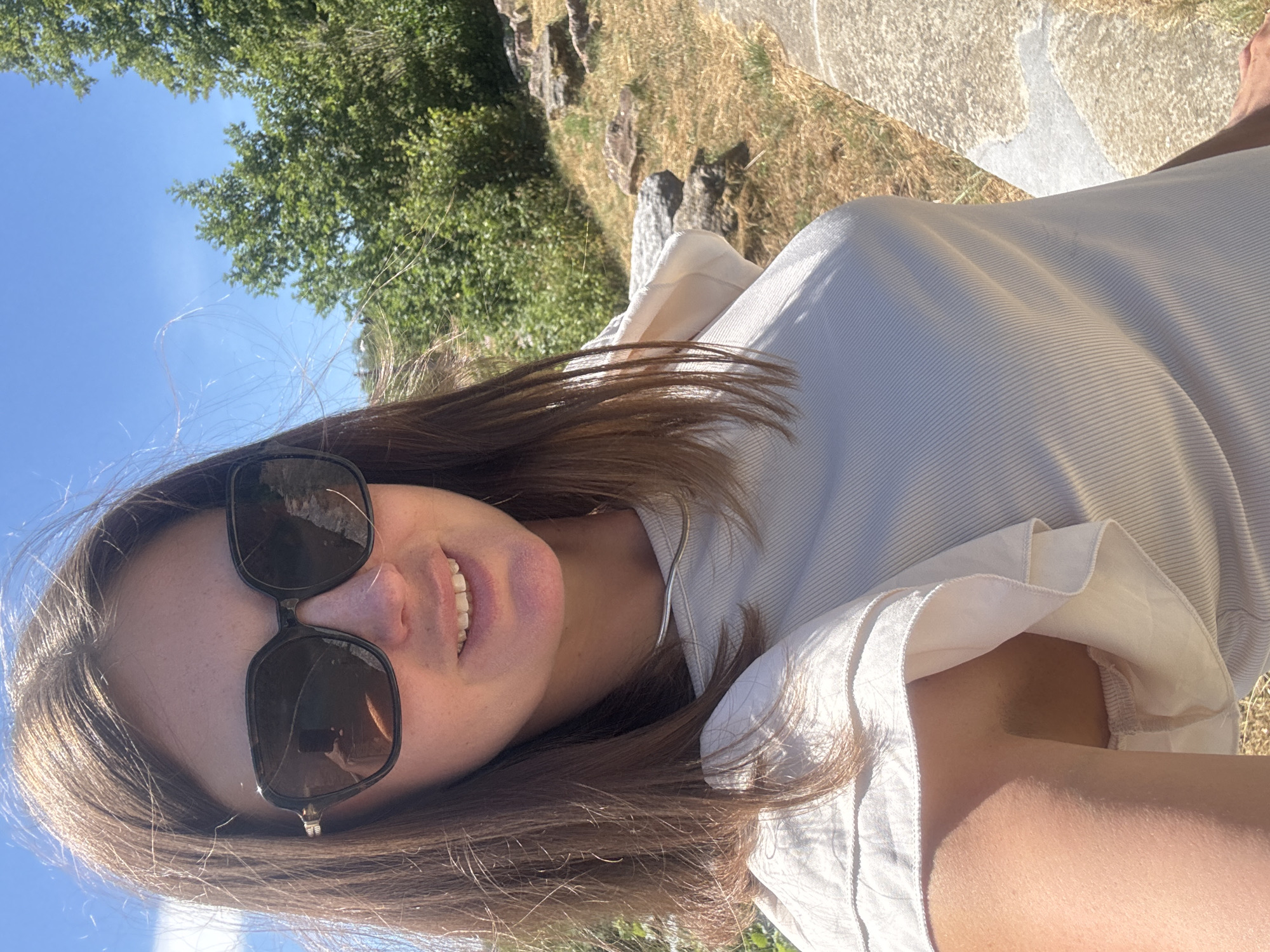A woman wearing large, dark sunglasses and a light-colored ribbed top takes a selfie outdoors on a sunny day. She has shoulder-length brown hair that is partially windswept. Her top has ruffled short sleeves. She is smiling, revealing her teeth. The background consists of green trees, a bright blue sky, and rocky terrain with dry grass. There is a pathway or wall made of stone to the right of the frame. The image appears to be taken at an outdoor location, possibly a park or a historical site.