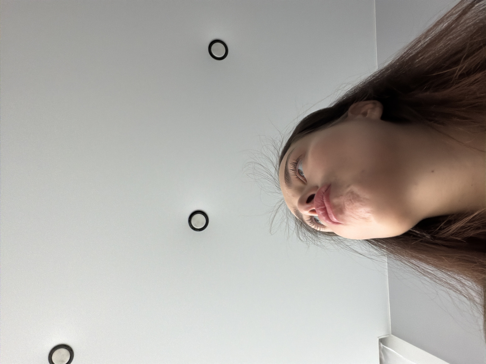 The image is taken from a low angle, looking up at a woman with long brown hair. Her face is positioned towards the upper right corner of the frame. Her lips are slightly pursed. The background consists of a white ceiling with three recessed light fixtures, appearing as black circles with white centers. The photograph seems to be taken indoors in the city of Молодёжный, Russia.