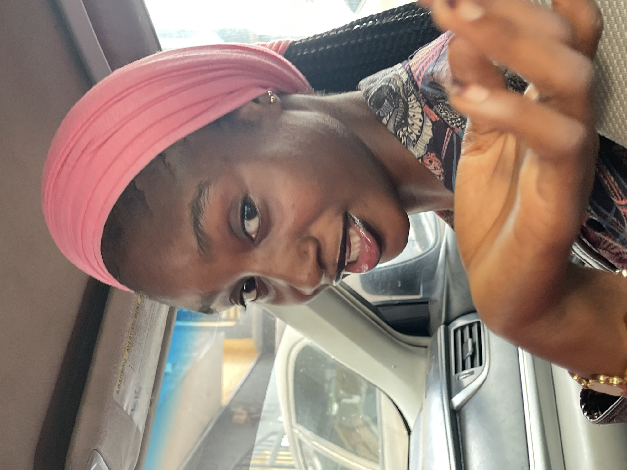 The image shows a young Black woman in the passenger seat of a vehicle, possibly in Abuja, Nigeria. She is wearing a pink headscarf and a patterned shirt. Her right hand is extended towards the camera, and she is looking directly at it with a playful expression. The interior of the car is visible, including the dashboard and a portion of another vehicle through the window. The overall lighting suggests it is daytime.