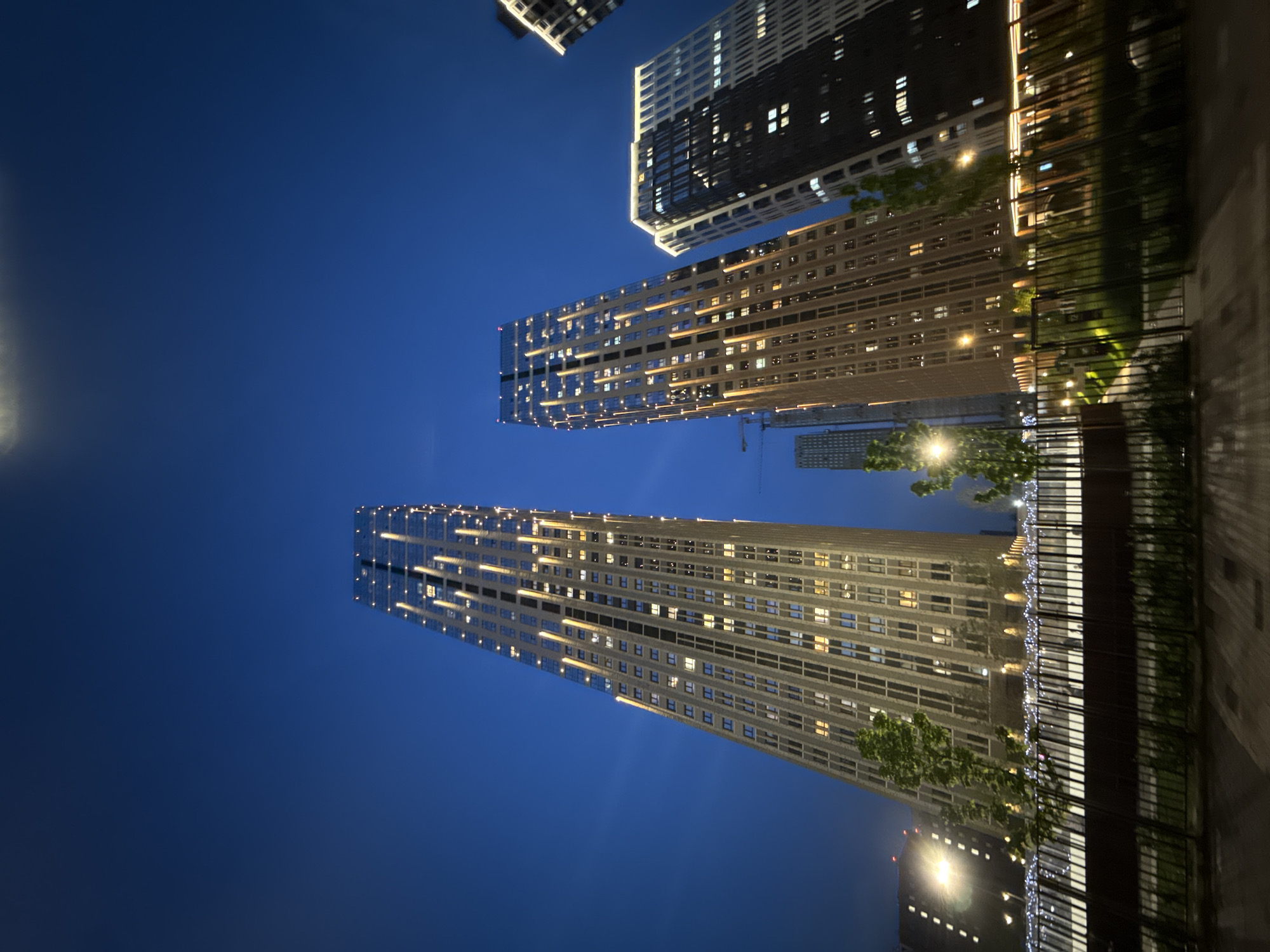 The image shows a view of several buildings in Moscow, Russia, taken at night. The sky is dark blue.
There are four buildings in the view. The two buildings closest to the bottom of the frame are illuminated with a repeating pattern of lights. The building closest to the top-center of the frame is also tall and lit with many lights. The fourth building is smaller and its lights are less visible in the distance.
In the lower-right corner, there is a dark, blurred object that looks like a fence or wall. There are trees along the right edge of the image and a bright light can be seen in the distance, possibly street lights.