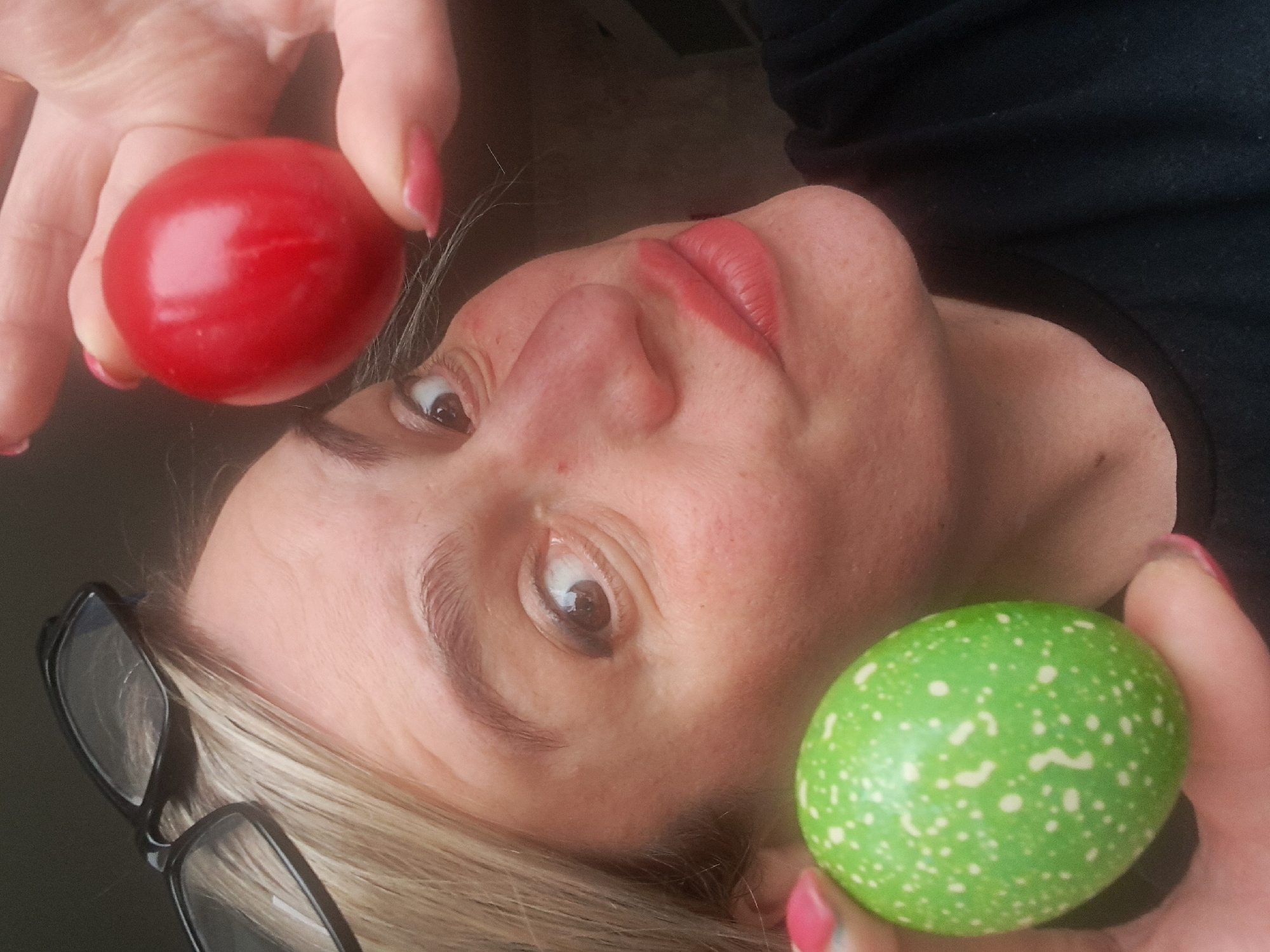 A woman is holding a red ball in her left hand and a green ball with white spots in her right hand. Her face is positioned between the two balls, and she is looking towards the viewer. A pair of black glasses rests on the left side of her head. Her hair is blonde. She has lipstick on. The background is dark. The location is not discernible.