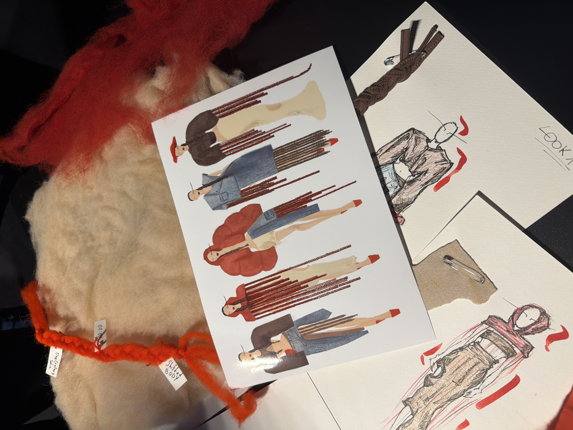 The image depicts a collection of fashion-related materials arranged on a surface. There is a printed card featuring illustrations of figures wearing clothing designs, with long braided extensions. There are also hand-drawn sketches of fashion designs on sheets of paper, some of which are held together with safety pins. Additionally, there are textile materials, including clumps of red and off-white fibers or wool. Small tags with handwritten text are attached to the red fibers. The scene appears to be a fashion designer's workspace. There is no explicit indication of the location being Sofia, Bulgaria, but it could be an interior space in that city.