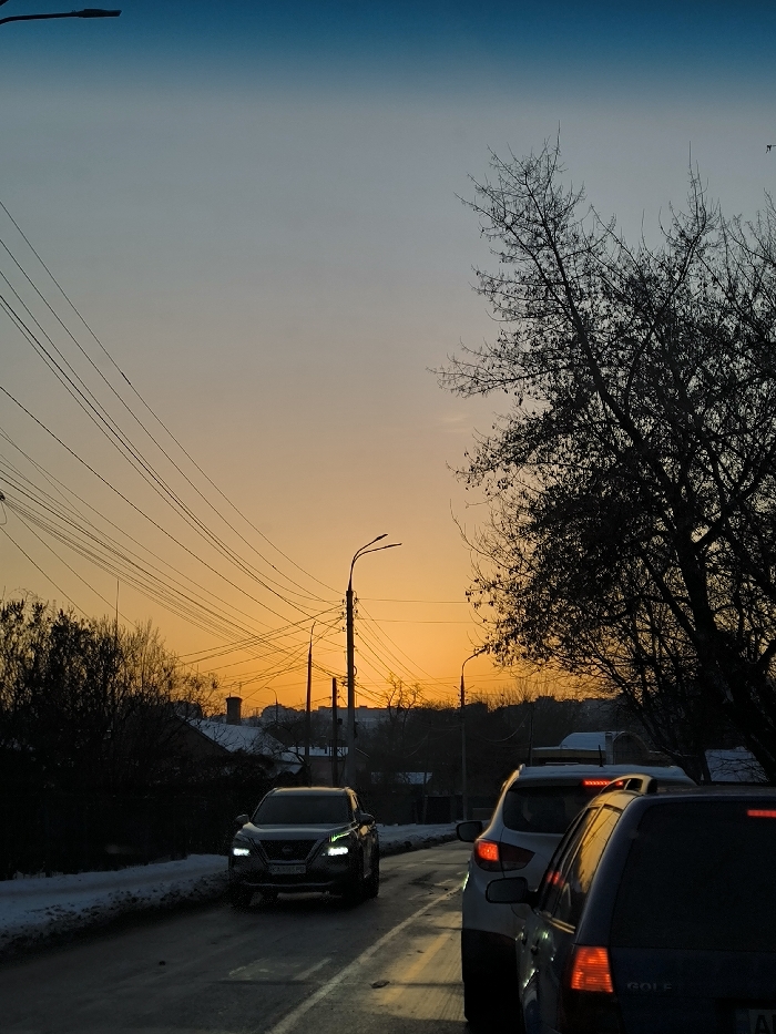 The image depicts a street scene in Vinnytsia, Ukraine, at dawn or dusk during winter. The sky transitions from a deep blue at the top to a vibrant orange-yellow at the horizon. The asphalt road in the foreground appears wet or icy, with patches of snow visible along its left edge.
Vehicles are present on the road. A dark-colored Nissan Qashqai (or similar SUV model) with illuminated headlights is visible on the left side of the lane, appearing to drive towards the viewer. In the right foreground, partially visible, are the rear sections of two vehicles: a white SUV/hatchback and, behind it, a dark blue Volkswagen Golf wagon. Both exhibit illuminated red brake lights, indicating they are either stopped or decelerating.
Overhead, numerous black utility lines crisscross the frame, supported by several utility poles. A prominent streetlight with two arms extends from one of these poles in the mid-ground. Silhouetted against the bright horizon are the bare branches of deciduous trees and the distant rooftops of buildings, confirming an urban or suburban setting.