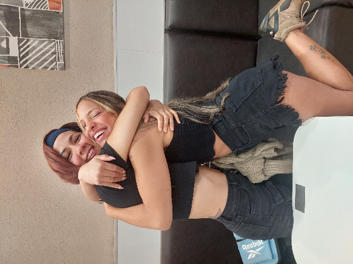 Two smiling women embracing each other. The woman on the left has short, auburn hair and wears a navy headband. The other woman has long, blonde-streaked hair. Both are wearing black tops and denim shorts. A brown couch and a beige sweater are visible in the background. One woman is lying on the couch. A piece of artwork adorns the wall. A tattoo is visible on the leg of the woman lying on the couch.