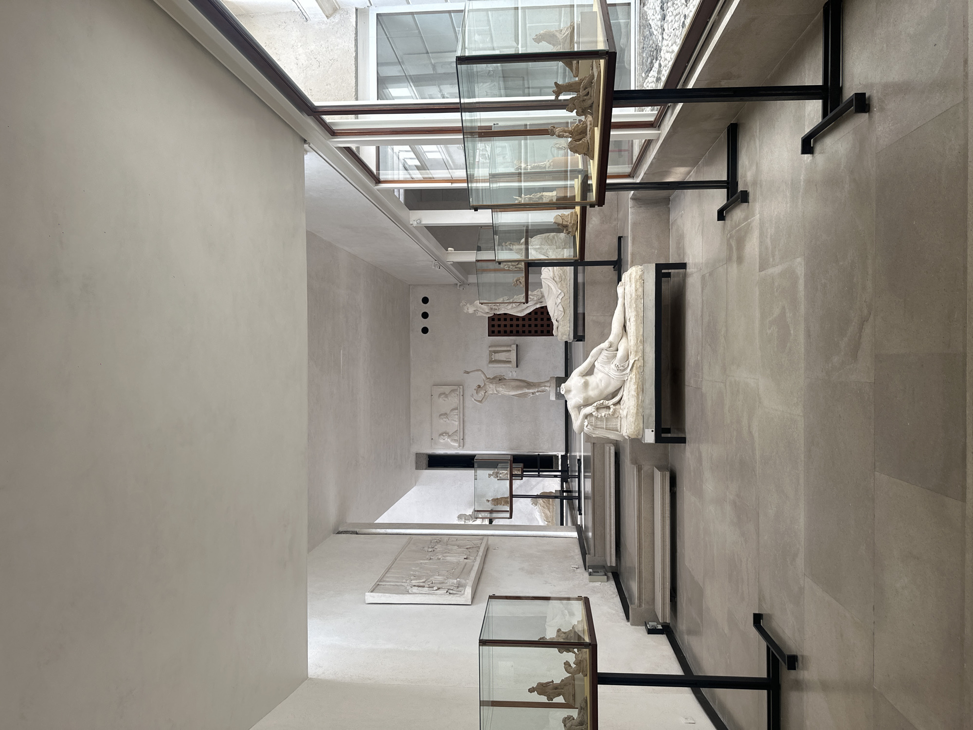 The image depicts the interior of a museum exhibit hall in Canicena, Italy, showcasing classical sculptures. The hall is spacious, with high ceilings and natural light filtering in from large windows on the right. The floor is tiled with large, pale grey stone slabs.
Dominating the foreground is a headless marble sculpture of a reclining nude figure. To the left, a similar marble sculpture of a nude figure is displayed on a raised platform. In the middle ground, a set of grey stone steps leads up to a slightly elevated area where more sculptures are displayed. One prominent sculpture on this level is a standing, draped female figure.
Several glass display cases on black metal stands are positioned throughout the hall, containing smaller sculptures and artifacts. These cases are illuminated, drawing attention to their contents. In the background, more relief sculptures are mounted on the white walls, and various statues are visible. The overall impression is one of an organized and well-lit exhibition space dedicated to classical art. The environment appears clean and well-maintained. No people are visible in the image.