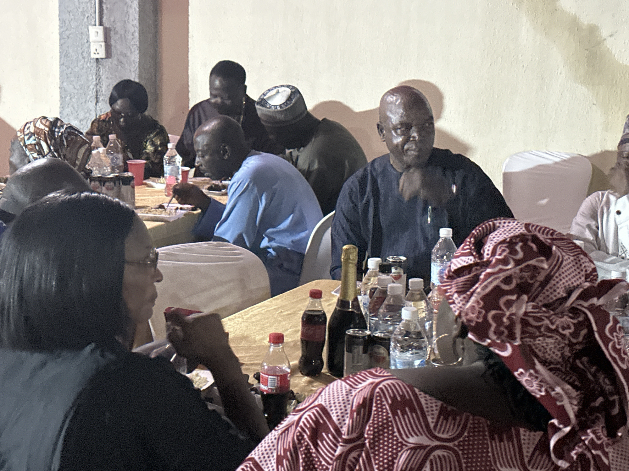 A gathering of people at a table in a room in Narayi, Nigeria. The table is covered with a tan cloth and has several bottles of water, Coca-Cola, and a bottle of champagne. People are sitting around the table, some eating and some talking. One woman in the foreground is wearing a red and white patterned headscarf. Another woman is wearing glasses and a black shirt. Several men are wearing traditional caps. The walls of the room are painted off-white.