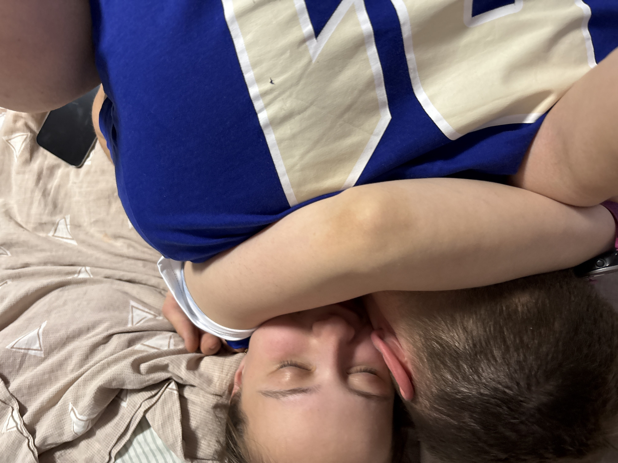 A close-up shot shows a woman with her eyes closed, lying on patterned bedding, possibly a bed. A person's arm is near her neck. Part of a blue shirt with cream-colored details is visible. A phone is nearby. The scene does not provide information to verify its location in the city of Прикамский, Russia.