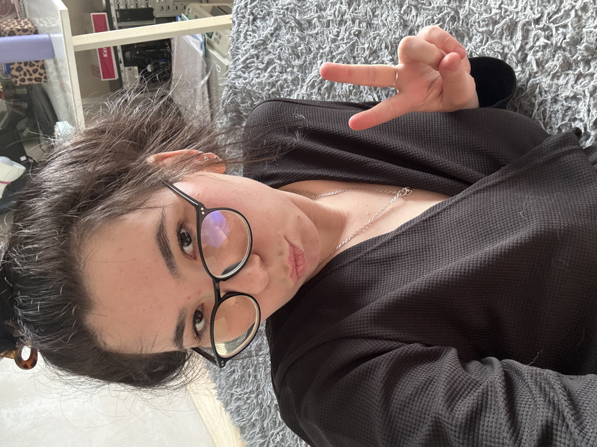 A woman with brown hair pulled back in a bun and wearing black-framed glasses is lying on a gray shag carpet. She is wearing a black, textured long-sleeved shirt and a silver necklace. Her right hand is raised, forming a "peace" sign. The background includes a partially visible white shelf unit and other miscellaneous items. The location is not possible to determine from the information in the image.