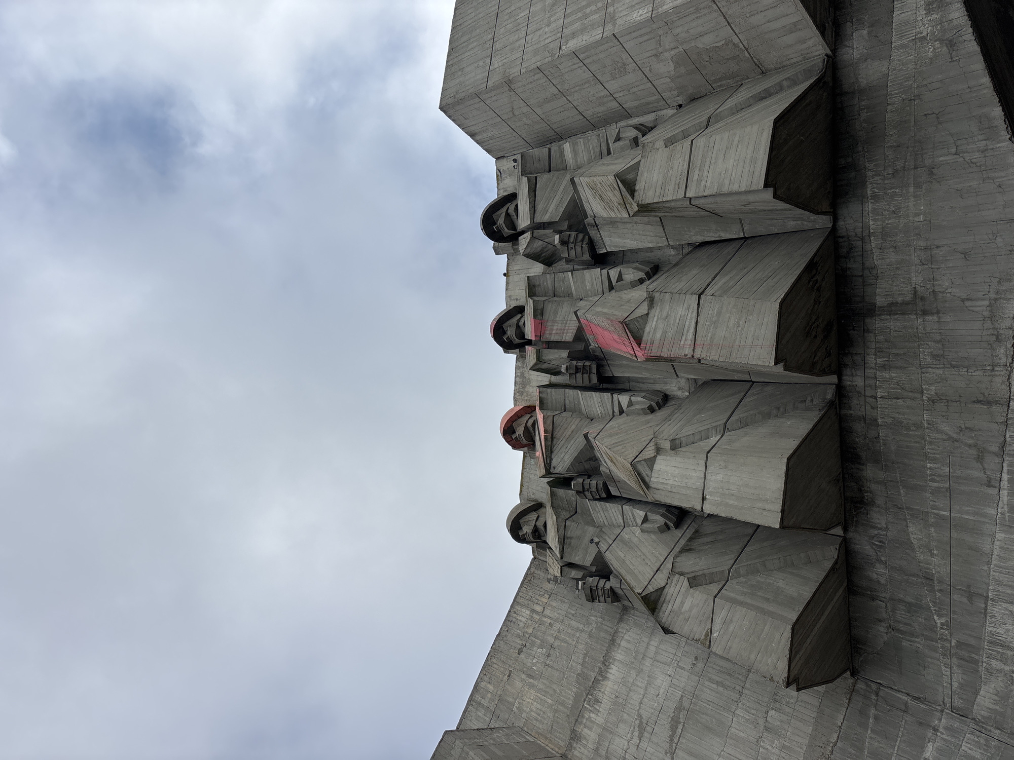 The image depicts a section of the Monument of the Bulgarian-Soviet Friendship in Varna, Bulgaria. The view is angled upwards, showing a concrete structure with reliefs of stylized figures wearing helmets. Some areas show red paint, indicating graffiti or vandalism. The background is a cloudy sky. The monument is constructed from large concrete blocks, giving it a brutalist architectural style.