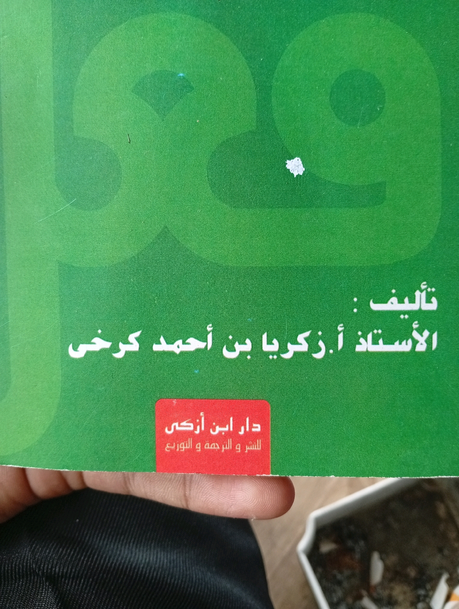 The image shows the cover of a book with Arabic text on a green background. The text appears to be the title and author of the book. There is also a red rectangular label with Arabic text on it, likely the publisher's name. A person's hand is visible at the bottom of the image, along with a white object that appears to be a container with some debris inside. The background is a textured green, likely the book cover itself. There is no visible indication of the city of Tasikmalaya, Indonesia, or any other specific location within the image.
