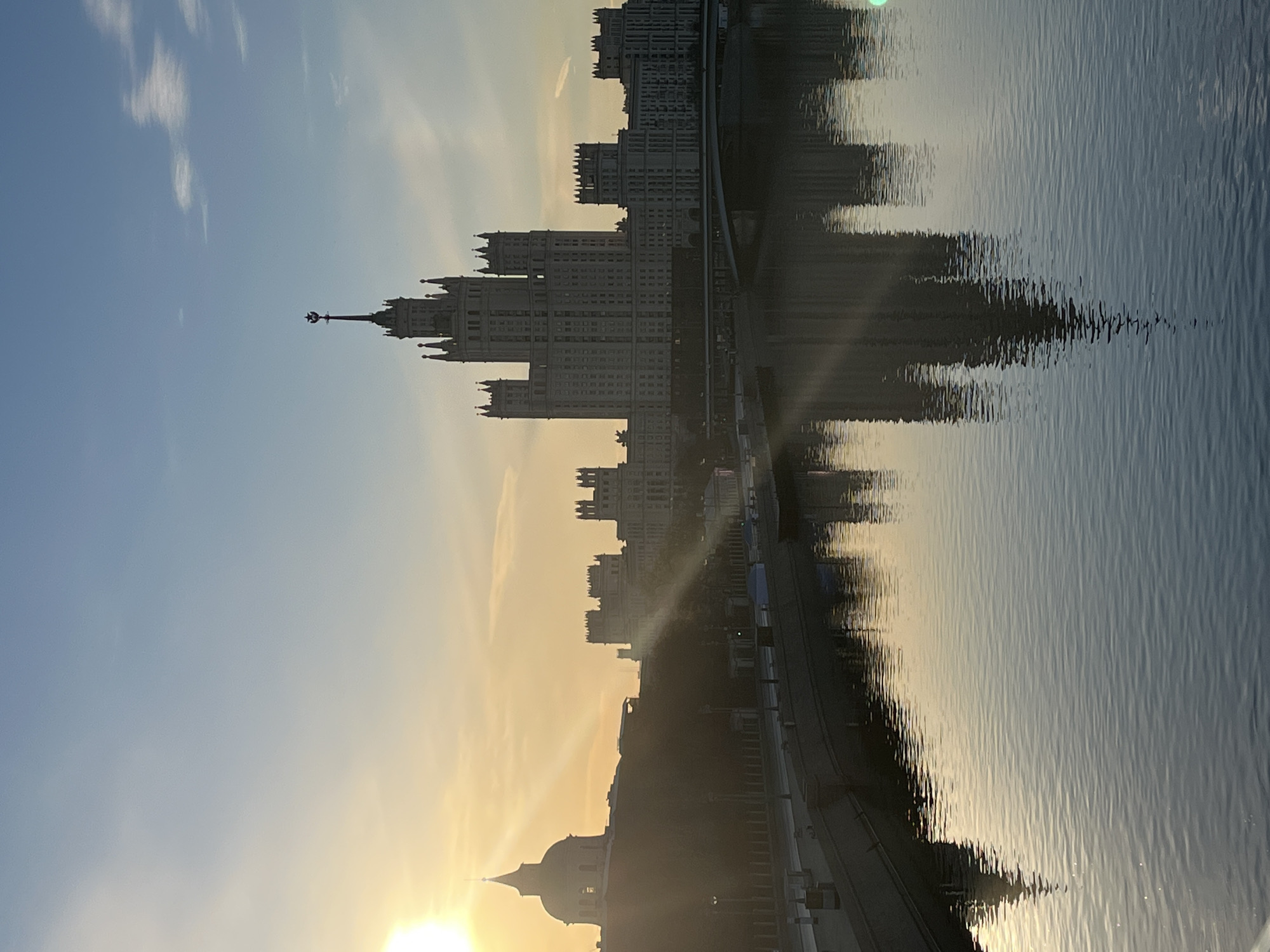 The image captures a scene in Moscow, Russia, at sunset. The dominant subject is one of the Seven Sisters skyscrapers, a prime example of Stalinist architecture, reflected in the water. The sun is low on the horizon, casting a warm glow. To the left, the sky is a gradient of blue and yellow, fading into the golden light near the horizon. The water's surface is ruffled with waves, distorting the reflection of the building. A road or walkway runs along the edge of the water, partially obscured by shadows.
