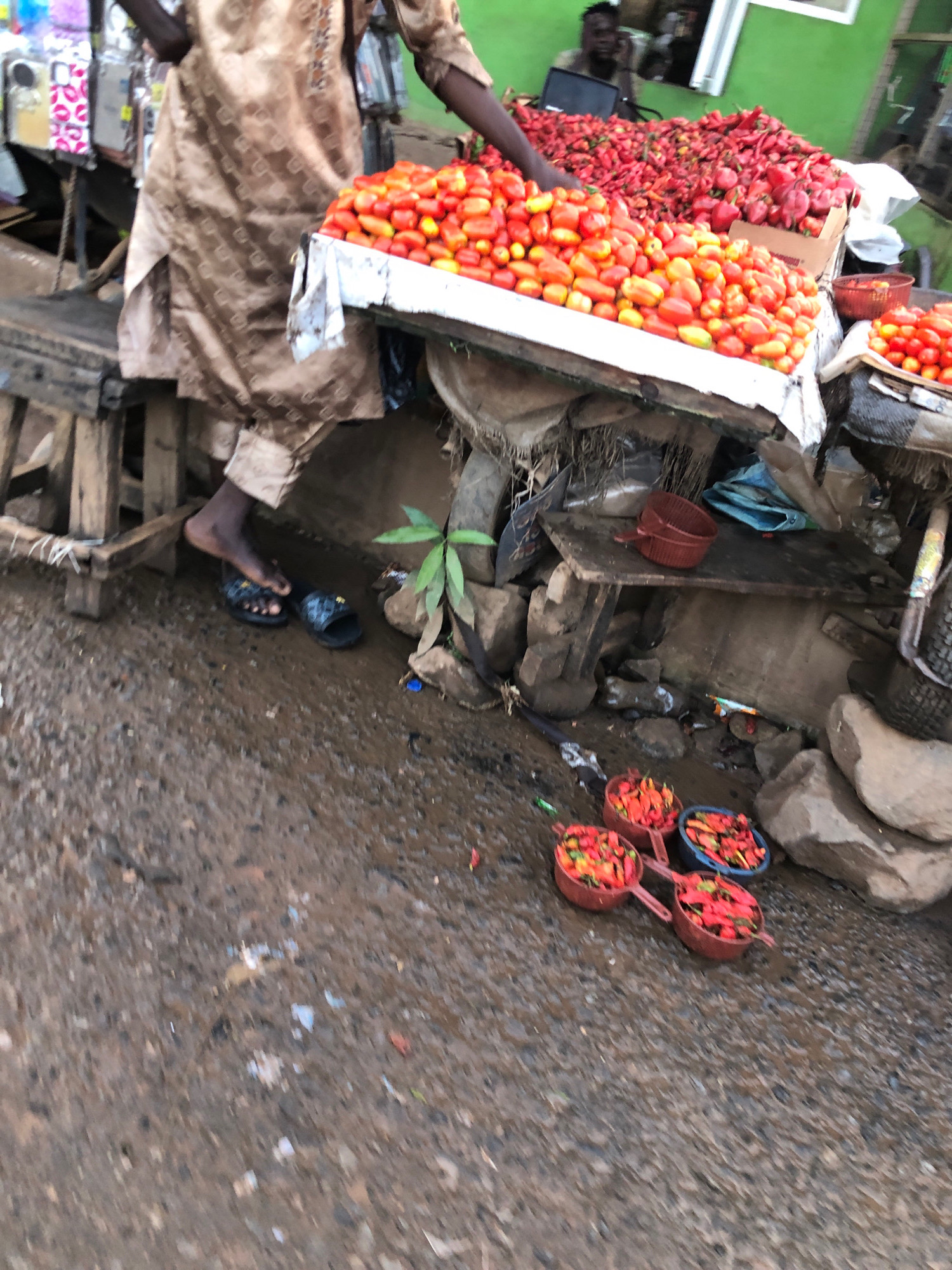 The image depicts a market scene at Aketu-Oja, Nigeria. A person, partially visible from the waist down, is standing next to a display of produce. They are wearing a patterned brown garment and dark sandals. In front of them, a wooden stall is laden with a large quantity of red tomatoes. Behind the tomatoes, a pile of red chili peppers is visible. To the right of the tomatoes, there are several red plastic bowls, some containing chili peppers, and a blue bowl also filled with chili peppers. The ground is wet and appears to be made of dirt or gravel, with some debris scattered around. The background is blurred but suggests a marketplace setting with structures and other vendors.