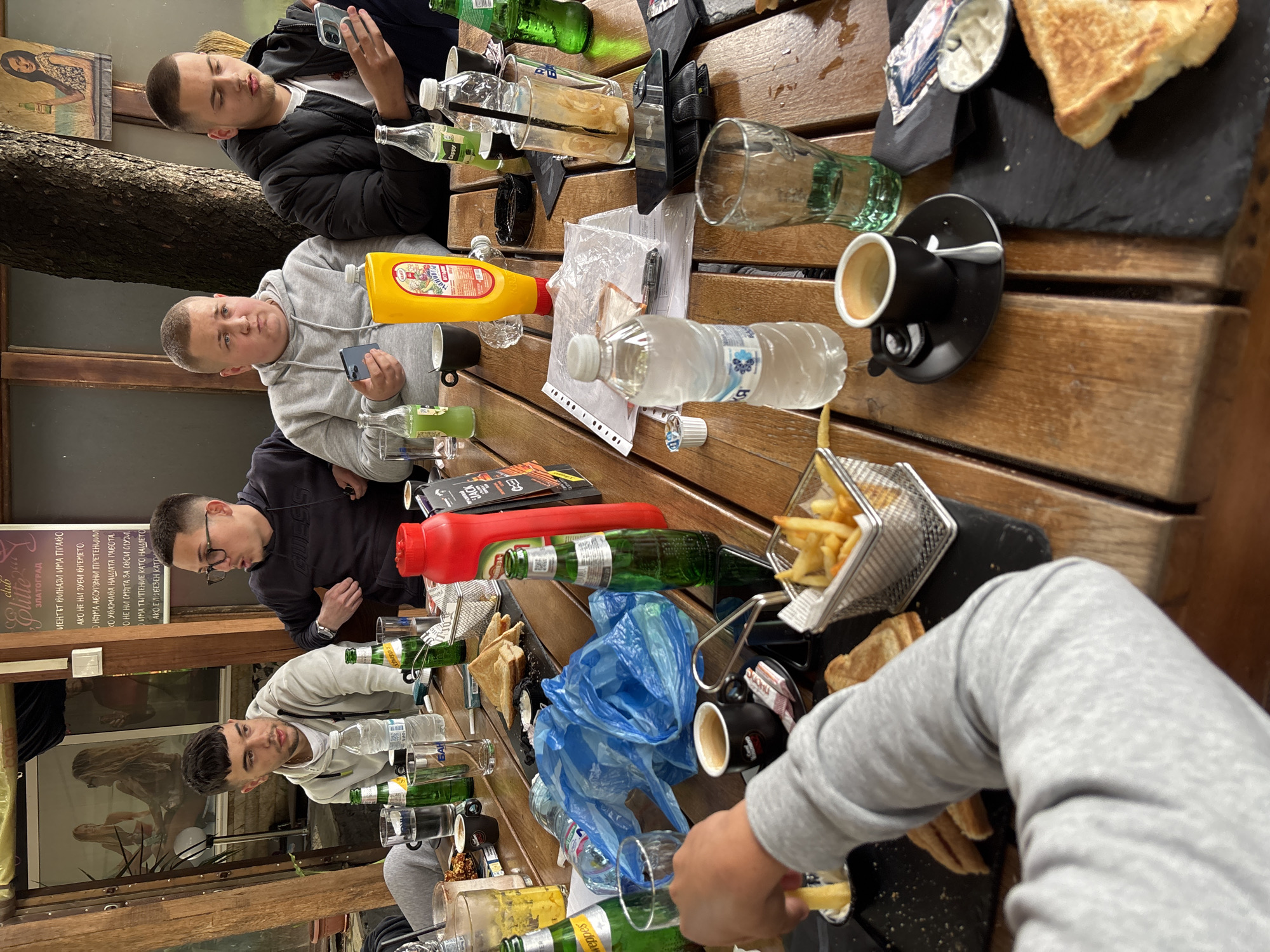 The image captures an outdoor dining scene in Zlatograd, Bulgaria, potentially at a cafe or restaurant called "Elite ЗЛАТОГРАД". Several young men are seated around a wooden table laden with food and drinks. The meal consists of french fries, sandwiches, and beverages in glass and plastic bottles. A yellow bottle of mustard is also present. Some individuals are interacting with smartphones. A blue plastic bag and personal items like wallets are scattered on the table. A sign is visible in the background with text related to the establishment’s service philosophy.