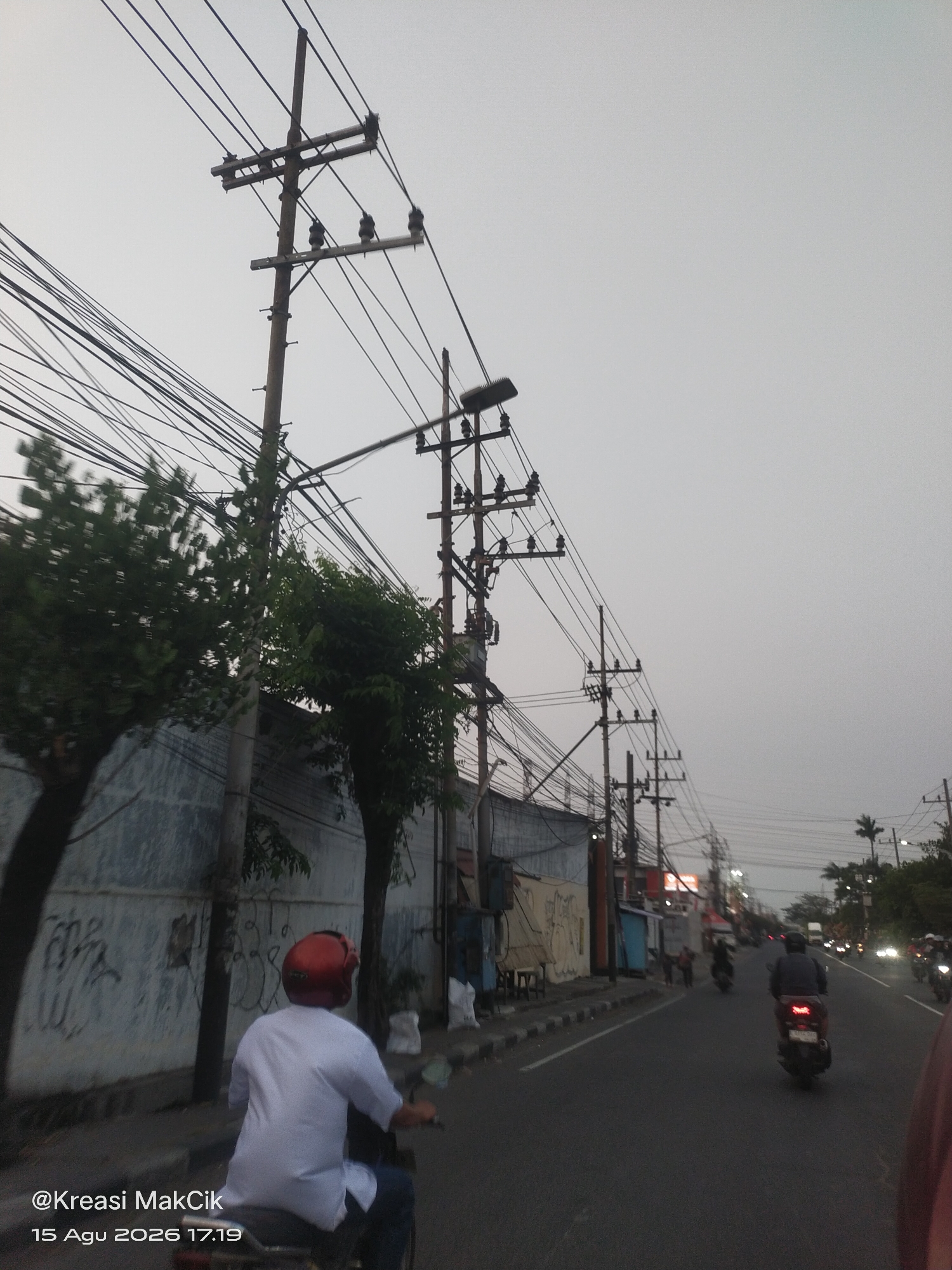 A perspective shot from a motorcycle captures a street scene in Surabaya, Indonesia, on August 15, 2026, at 17:19. Overhead, a dense network of power lines crisscrosses the sky, supported by multiple utility poles. A person wearing a white shirt and a red helmet is riding a motorcycle in the foreground, their back to the camera. The street is lined with buildings and trees on the left, with some graffiti visible on a wall. Several other motorcycles and a truck are visible on the road ahead. The sky is overcast, and the overall lighting suggests it is late afternoon or early evening. The watermark "@Kreasi MakCik" and the date are superimposed on the lower left of the image.
