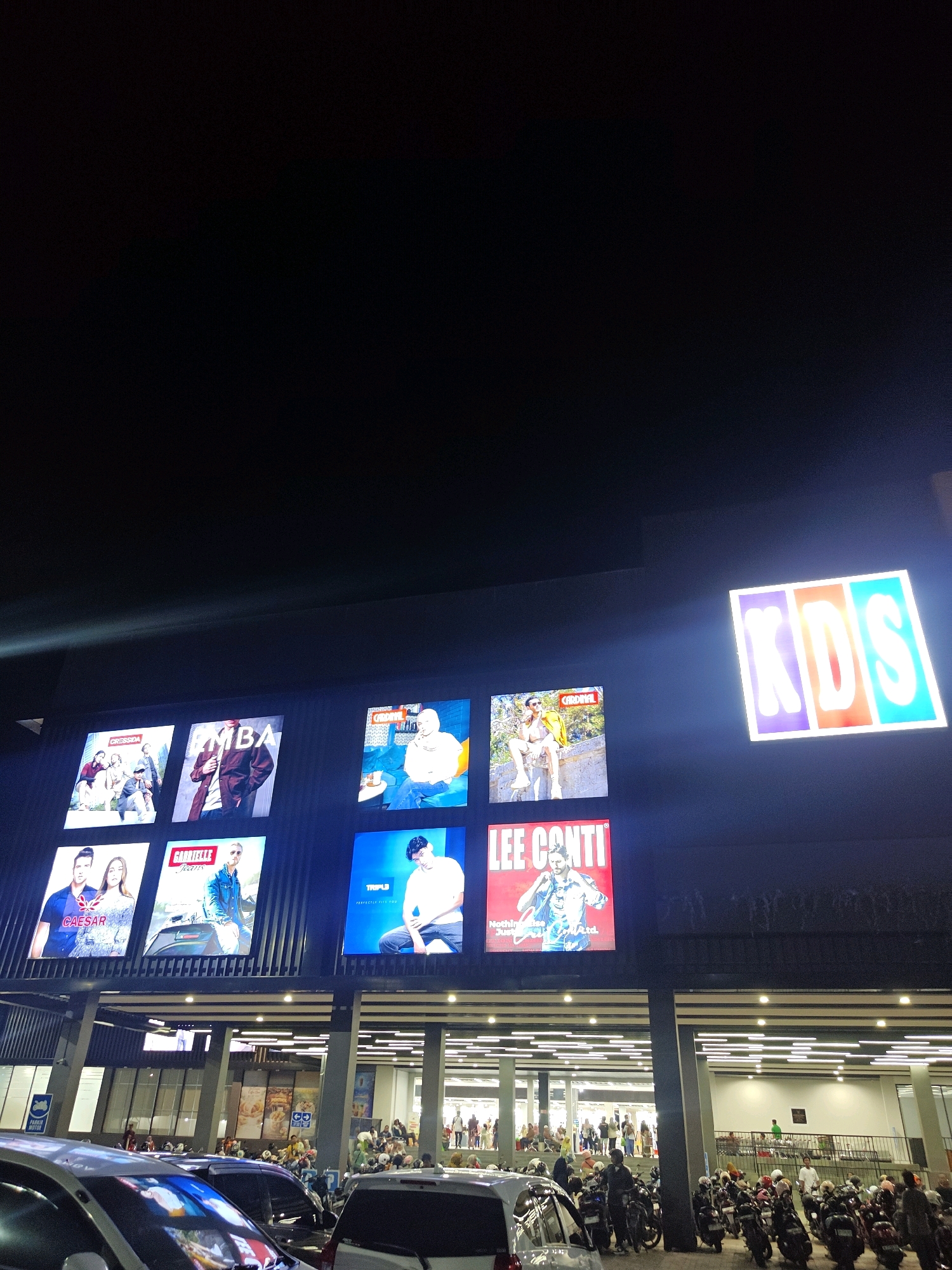 The image shows the exterior of the KDS department store in Situbondo, Indonesia at night. A large, illuminated sign with the letters "KDS" in block capitals is visible on the building's facade. Below the sign, several large screens display advertisements. One screen shows a man in a white shirt and jeans. Another displays an advertisement for "LEE CONTI" with a picture of a man in a denim jacket. Other screens show advertisements for fashion brands and possibly movie posters. The building has a modern architectural design with a dark facade and a series of horizontal lights. In front of the store, a parking lot is filled with motorcycles and a few cars. People are seen entering and exiting the building, and some are gathered in the parking area. The overall atmosphere is that of a busy commercial area at night.