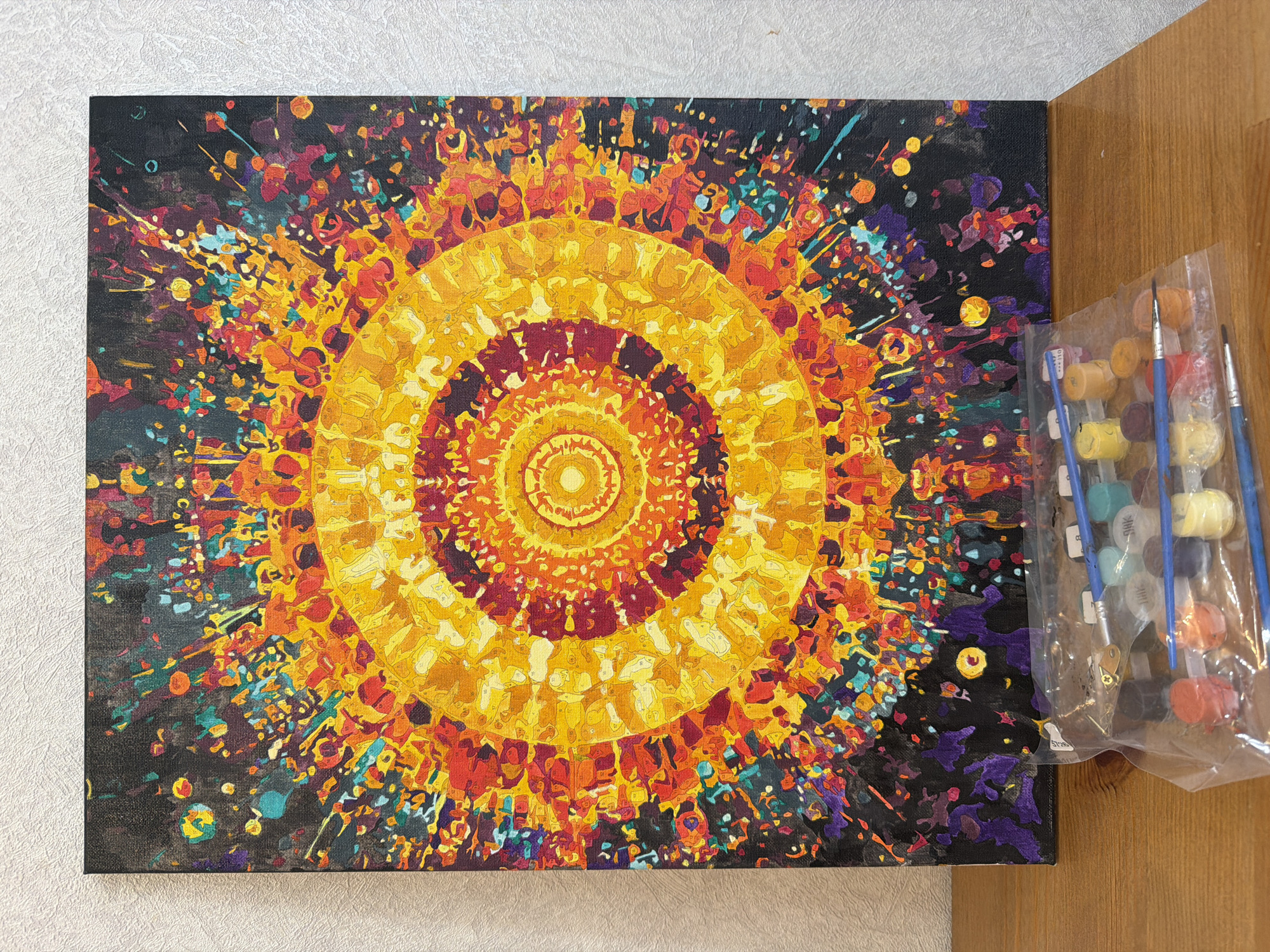 The image presents a close-up, high-angle view of a completed paint-by-numbers project depicting a vibrant, abstract circular design reminiscent of a sun or explosion. The artwork is on a canvas, predominantly featuring concentric rings of warm colors including yellow, orange, and red, set against a darker background filled with multicolored flecks and streaks. The canvas sits on a light-colored surface, possibly a tabletop. To the right of the canvas, a clear plastic bag contains multiple small paint pots with corresponding brushes, suggesting the materials used for the paint-by-numbers activity. The details present no indication of Myaglovo, Russia.