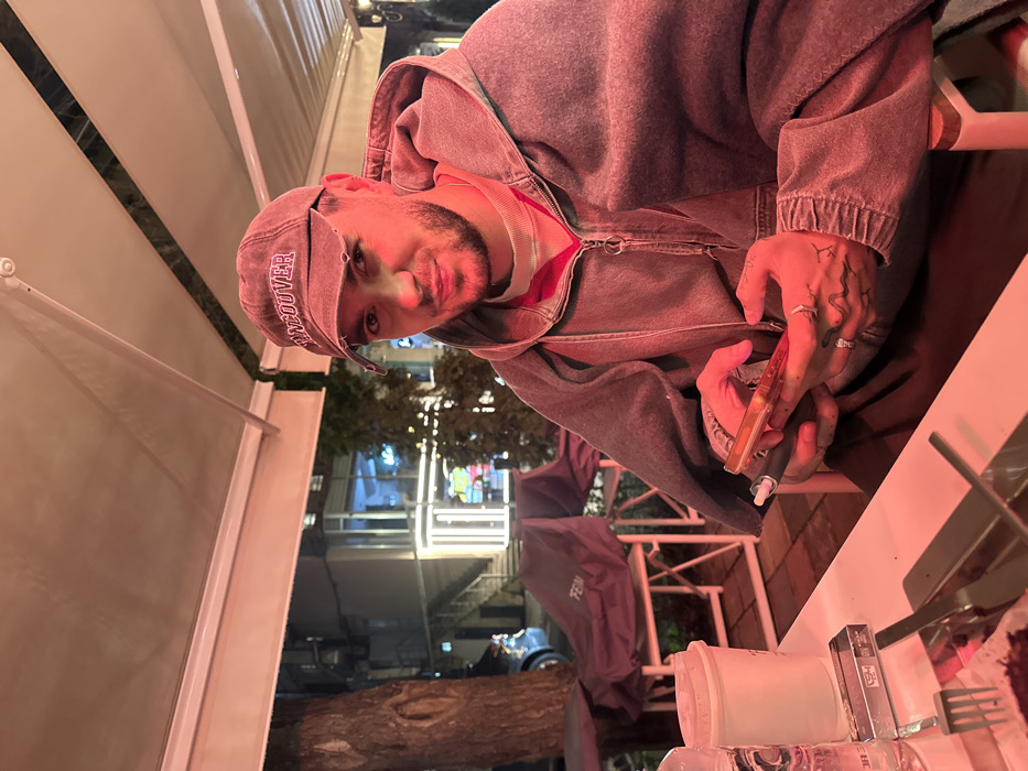 A young adult male is seated at an outdoor dining table, looking directly at the camera with a slight smirk. He wears a faded reddish-brown "VANCOUVER" baseball cap, a grey zip-up hoodie over a light-colored turtleneck, and has a short beard. Multiple rings adorn his left hand, which also displays a snake-like tattoo. He holds a gold-colored smartphone with an attached vape pen in both hands.
On the white table in front of him are a clear glass water bottle, a white disposable cup, a dark cigarette pack (likely "GM" or "Garni MM"), a fork and knife, and a plate containing dark-colored food. The scene is illuminated by a reddish artificial light, likely from an overhead heat lamp, under a light-colored retractable awning.
In the mid-ground, an outdoor seating area features chairs and tables covered with dark protective tarpaulins, one of which visibly reads "FERM." A tree trunk is to the left. The background reveals a street with a blue car, and modern buildings exhibiting illuminated windows and vertical strip lighting, indicative of an urban environment.
Based on the visible "Garni MM" cigarette brand and the "FERM" branding (consistent with Fermata Restaurant), the location is identified as an outdoor dining area in Yerevan, Armenia, likely Fermata Restaurant.