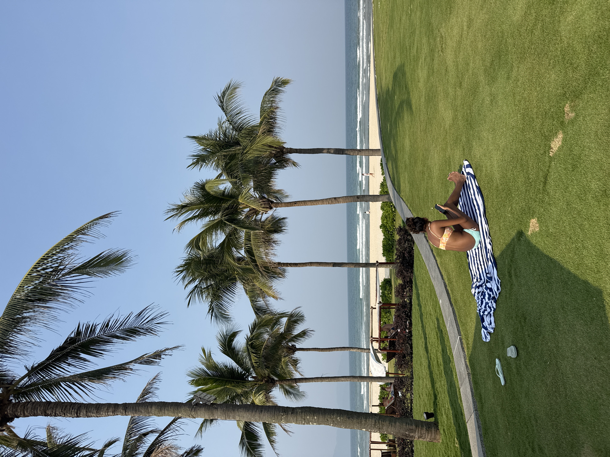 A woman is sitting on a striped beach towel on a grassy lawn in Đà Nẵng, Vietnam. She is wearing a bikini and using her phone. Palm trees are in the background, with the beach and ocean visible beyond. The grass appears to be well-maintained. Two pairs of flip-flops are near the towel. A walkway borders the grassy area. Seating under sun protection is visible in the background. The sky is clear and blue.