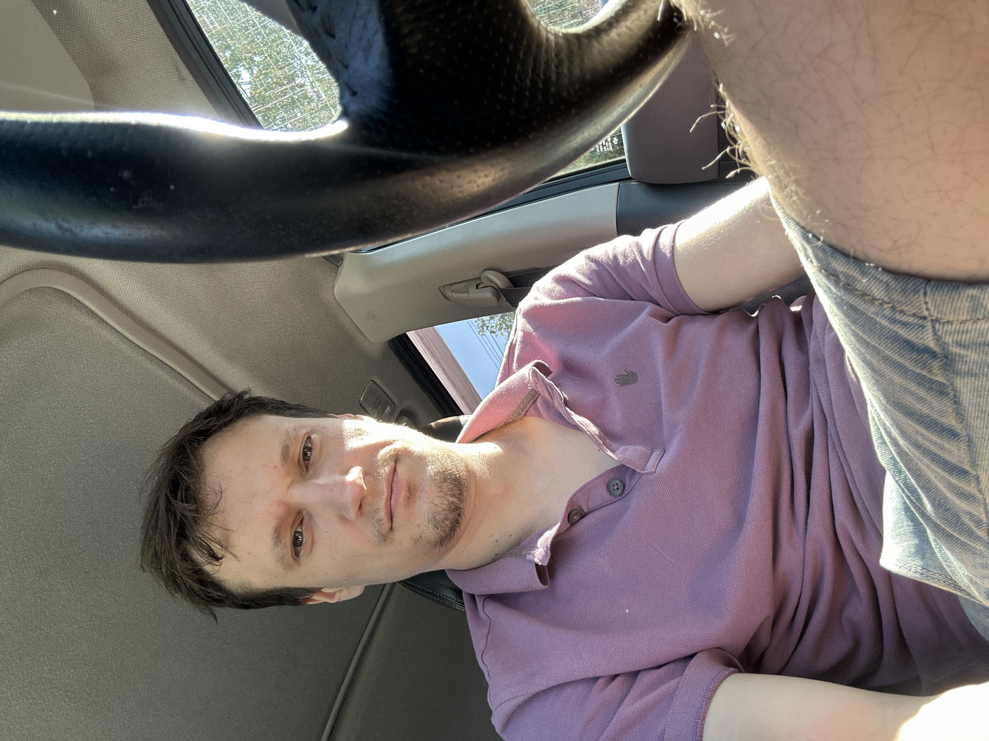 A man, appearing to be in his late 20s or early 30s, is captured in a selfie from the chest up. He is seated in the driver's seat of a vehicle, with the steering wheel prominently visible on the right side of the frame. He has light brown hair, brown eyes, and a light stubble on his chin and jawline. He is wearing a purple short-sleeved polo shirt and light blue denim shorts. The interior of the car is visible, including the headliner and a portion of the passenger side. Sunlight streams in from the right, illuminating the scene and casting shadows. The background outside the car window suggests an outdoor environment with trees and possibly a building. The image was taken in the city of Makiv, Ukraine.