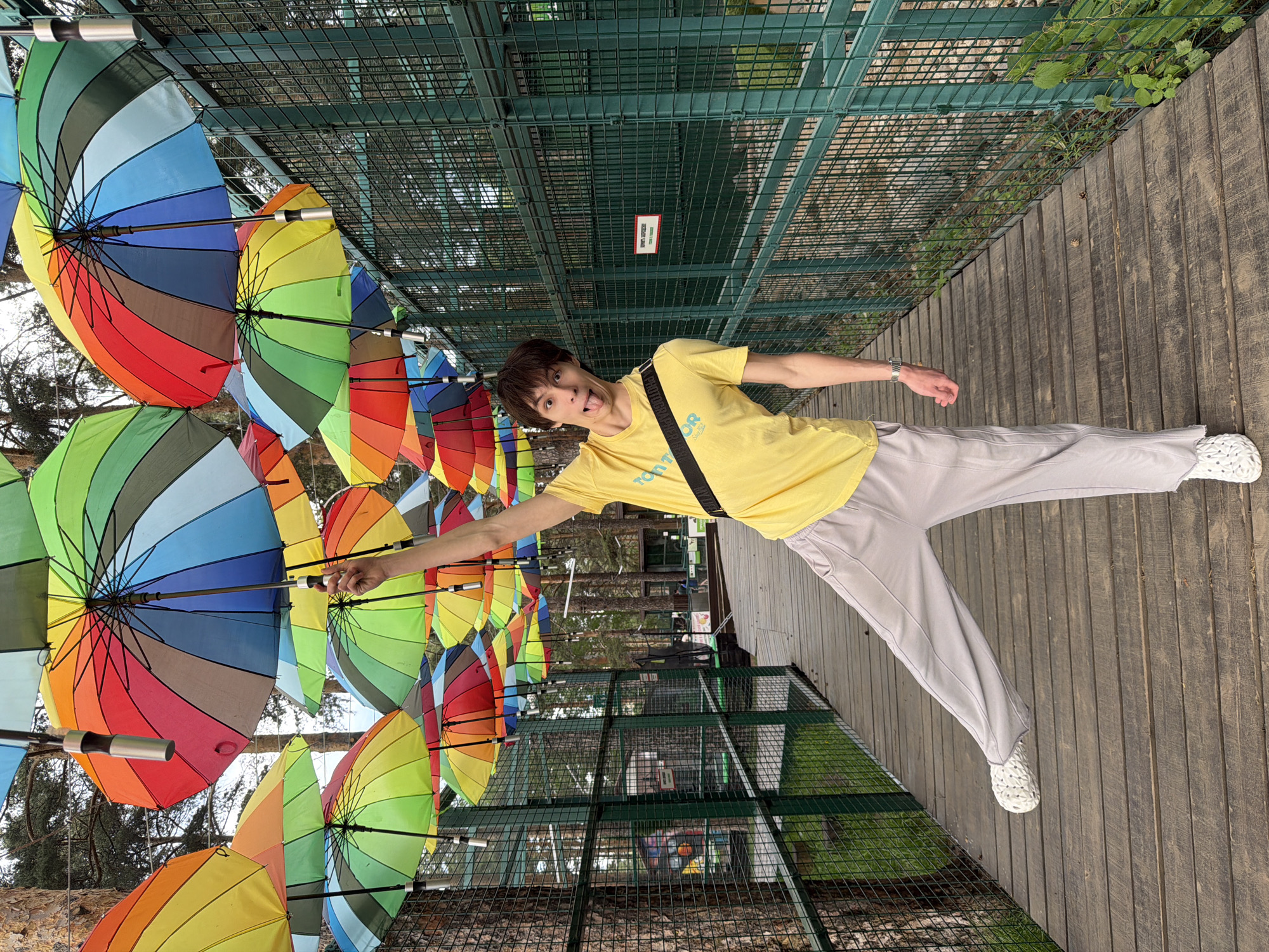 A young adult stands on a wooden boardwalk, extending one arm and holding an umbrella from a colorful array of open umbrellas suspended above. The location is Yekaterinburg, Russia. The individual wears a yellow t-shirt, light grey pants, and white clog-style shoes. The background includes a green, wire mesh fence and trees.