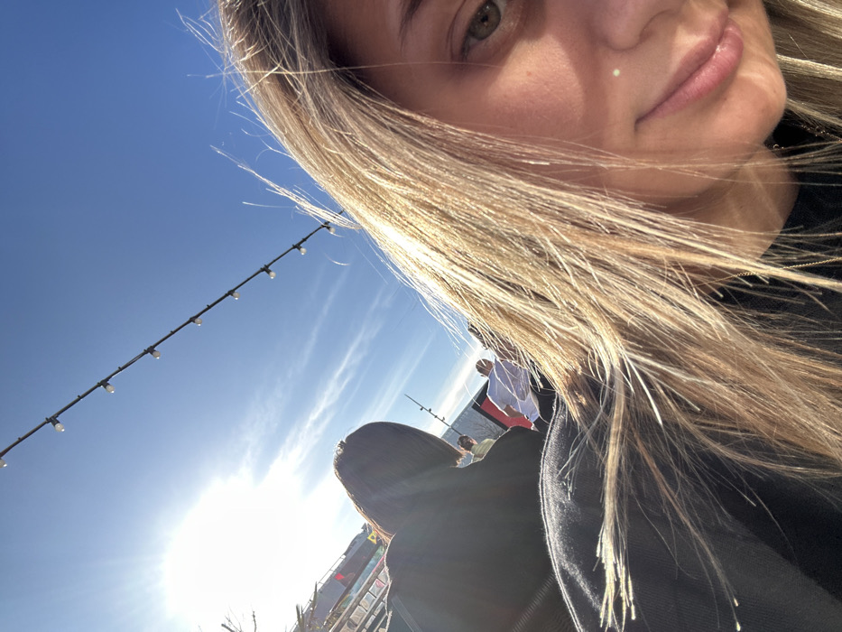 A selfie-style shot angled upwards. A fair-skinned woman with blonde hair occupies the upper right frame. Part of another individual's black-clothed back and head is visible in the lower center. In the background, a blue sky is dotted with light clouds and a string of lights is strung across the upper left. A bright, blurred sun creates intense glare.