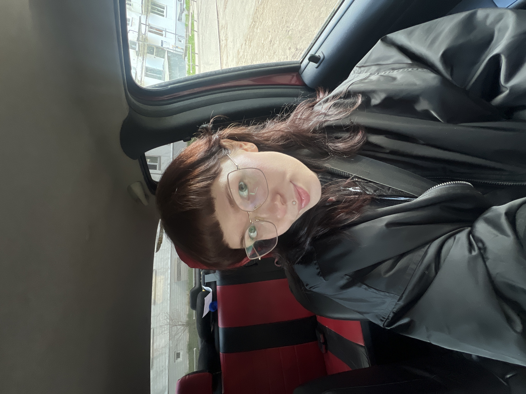 A person with reddish-brown hair and glasses is in the front seat of a car with red and black seats. The person is wearing a black jacket. Outside the car window, there is a multi-story building visible. The location is Chaikovsky, Russia.