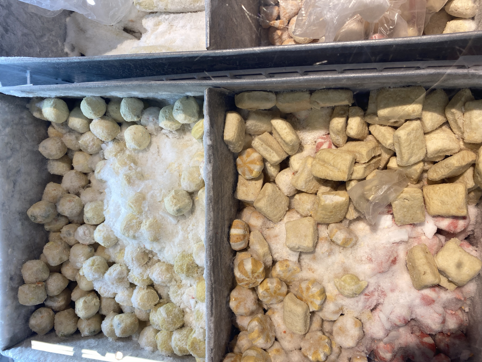 The image shows a close-up, top-down view of frozen food products displayed in a refrigerated display case, likely in a market or supermarket in Banda Aceh, Indonesia. The display case is divided into compartments.
The upper compartment contains spherical, pale yellow frozen food items, possibly fish balls or similar dumplings, covered in a layer of frost and ice. The items are irregular in shape and size, with some appearing slightly flattened.
The lower compartment displays a variety of frozen food items. On the left side of this compartment, there are small, round, pale yellow food items with orange stripes, resembling small buns or dumplings. Scattered among these are some cubic, pale yellow items and a layer of pink and white ice crystals, suggesting other frozen products might be underneath. To the right of these, there are more cubic, pale yellow frozen food items, appearing to be some kind of processed tofu or a similar product. Some of these cubes have a textured surface. In the far right of this lower compartment, partially visible plastic packaging with frozen food items can be seen.
The overall environment appears to be a commercial food display area, characterized by the metallic bins of the display case and the presence of frost and ice, indicating a cold storage environment. The lighting suggests overhead illumination, typical of a retail setting. No people are visible in the frame. The context of Banda Aceh, Indonesia, implies a tropical climate where refrigerated displays are essential for food preservation.