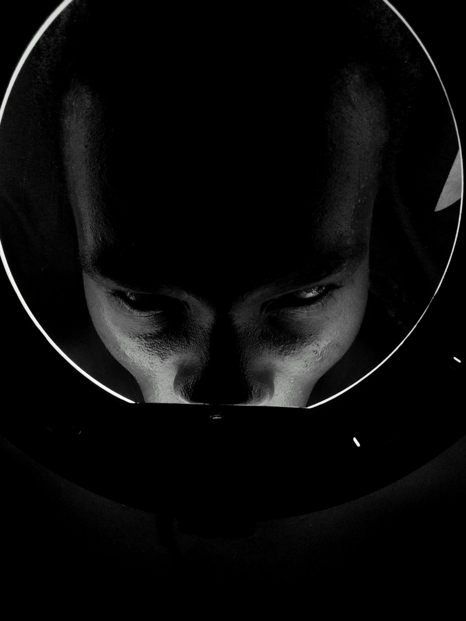 A black and white photograph depicts a person's face looking down into an unknown object. The person is positioned in the upper center of the image, surrounded by a circular white light. The lower half of the face, including the nose, mouth, and part of the chin, is visible. The eyes are directed downwards. The surrounding area is dark, with a large, dark circular object in the lower portion of the image. The location is Unknown, Unknown.