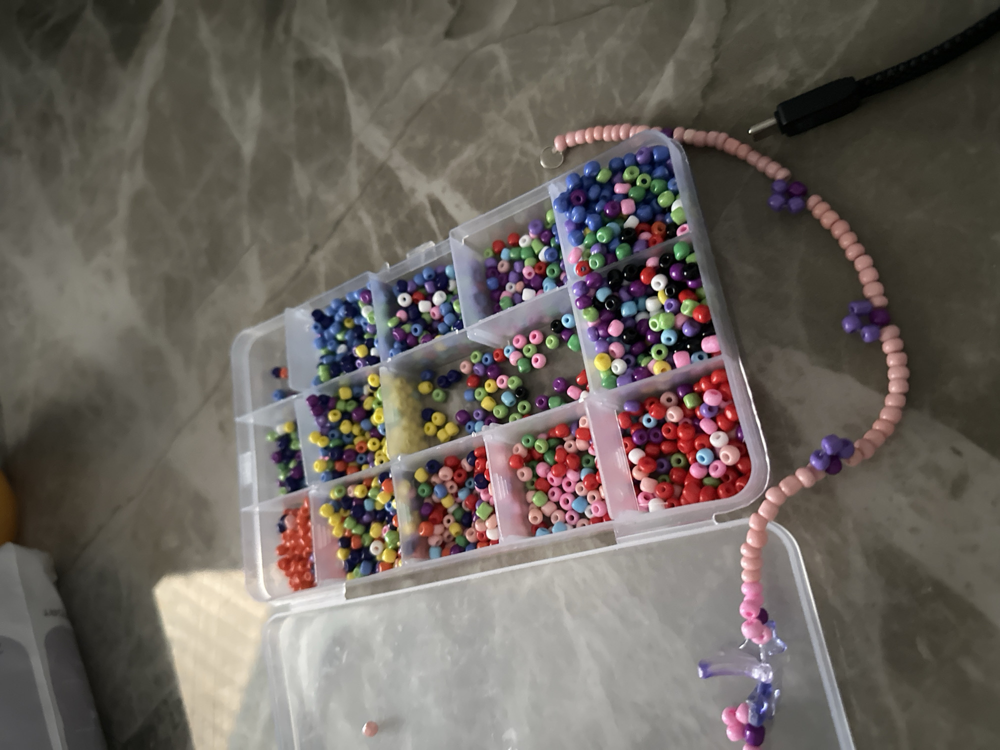 The image shows a plastic container with multiple compartments filled with colorful seed beads, mostly blue, green, red, and yellow. A beaded necklace made of pink beads and purple flower-shaped beads extends from the top right corner of the container. A black USB cable is located near the necklace. The background appears to be a grey surface. The image was taken indoors, presumably in Bac Nha Trang Ward, Vietnam, given the prompt's requirement.