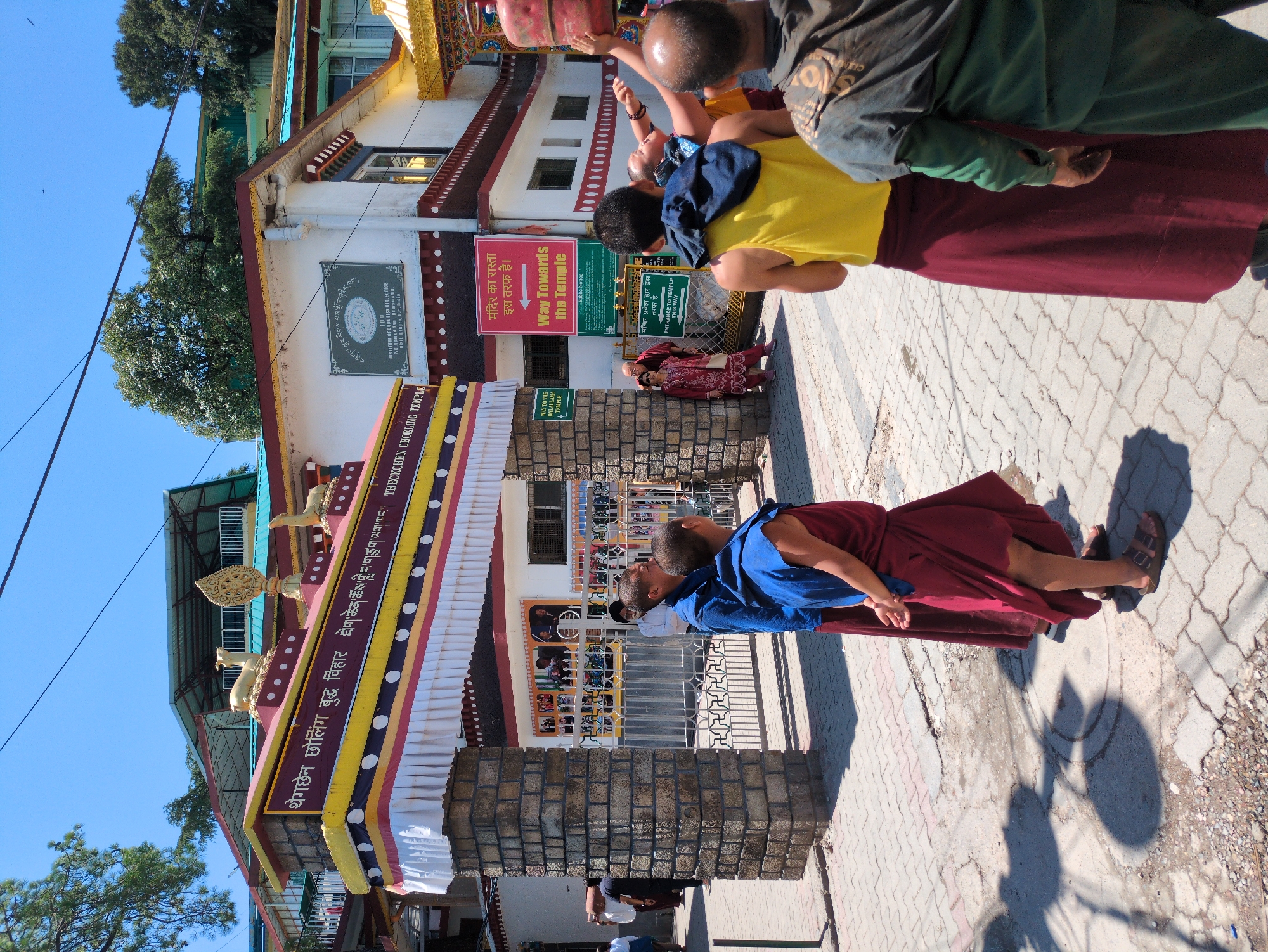 The image depicts the entrance to the THeCKCHEN CHOLING TEMPLE in Dharamshala, India. The architecture features a decorative archway with Tibetan inscriptions and ornate golden statues of mythical creatures. Several individuals are present, including monks in maroon and blue robes and other people in casual attire. Some are walking, while others are standing and observing. Two young monks in maroon robes are positioned in the foreground, facing away from the camera. A person in a yellow vest and maroon skirt is also in the foreground, with their back to the viewer. A sign with text in Hindi and English, "Way Towards The Temple," is visible on the right side of the entrance. The environment appears to be a street with paved pathways and buildings in the background, suggesting a bustling urban or semi-urban setting within Dharamshala, a town known for its Tibetan community and Buddhist monasteries. The sky is clear and blue, indicating a sunny day.