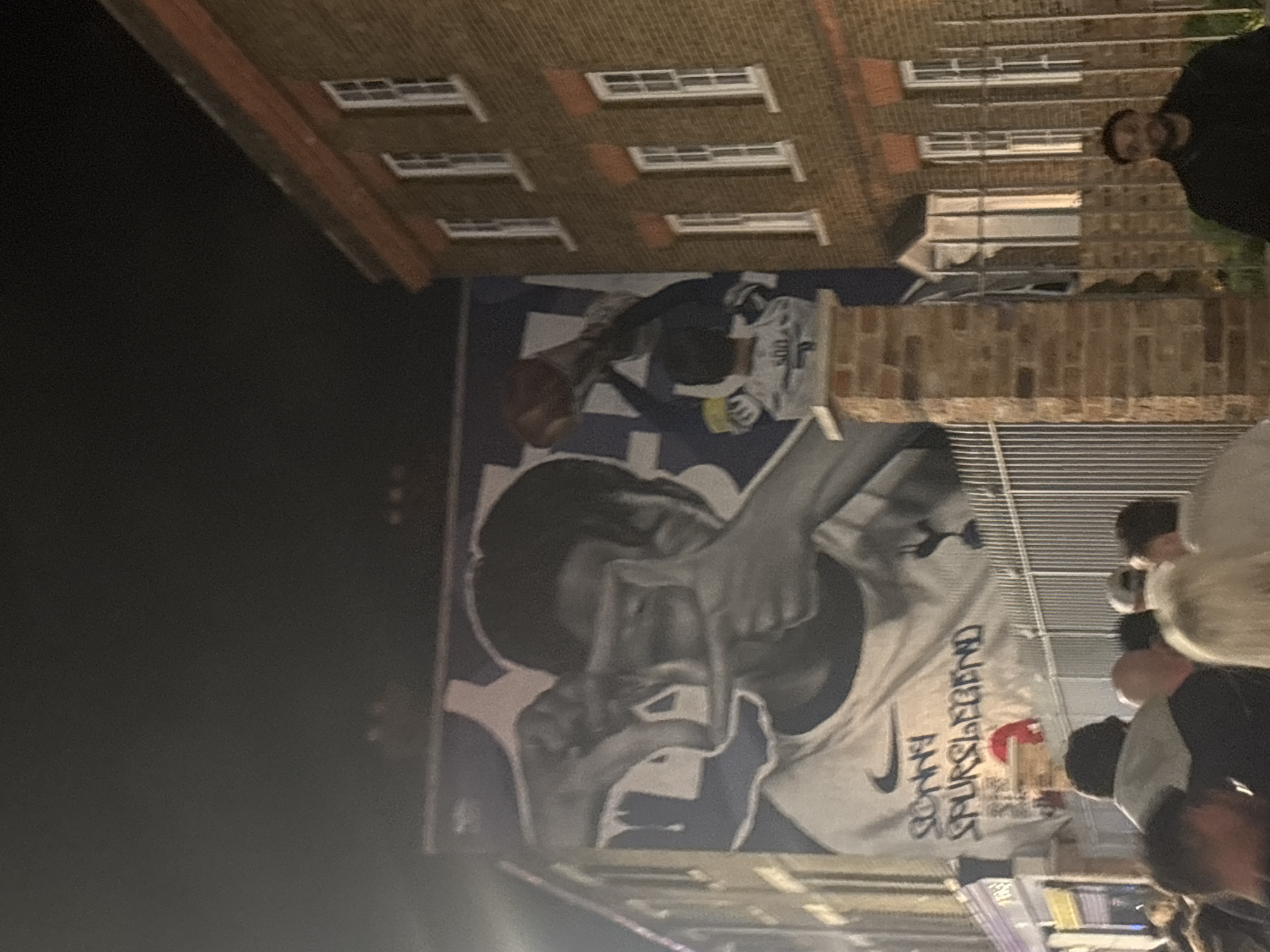 A mural depicting a person with text is on the side of a brick building. The text on the mural reads, "Sonny Spurs Legend." The location appears to be in London, United Kingdom. People are standing around a table in front of the mural. A man is visible in the background near the top right of the image.