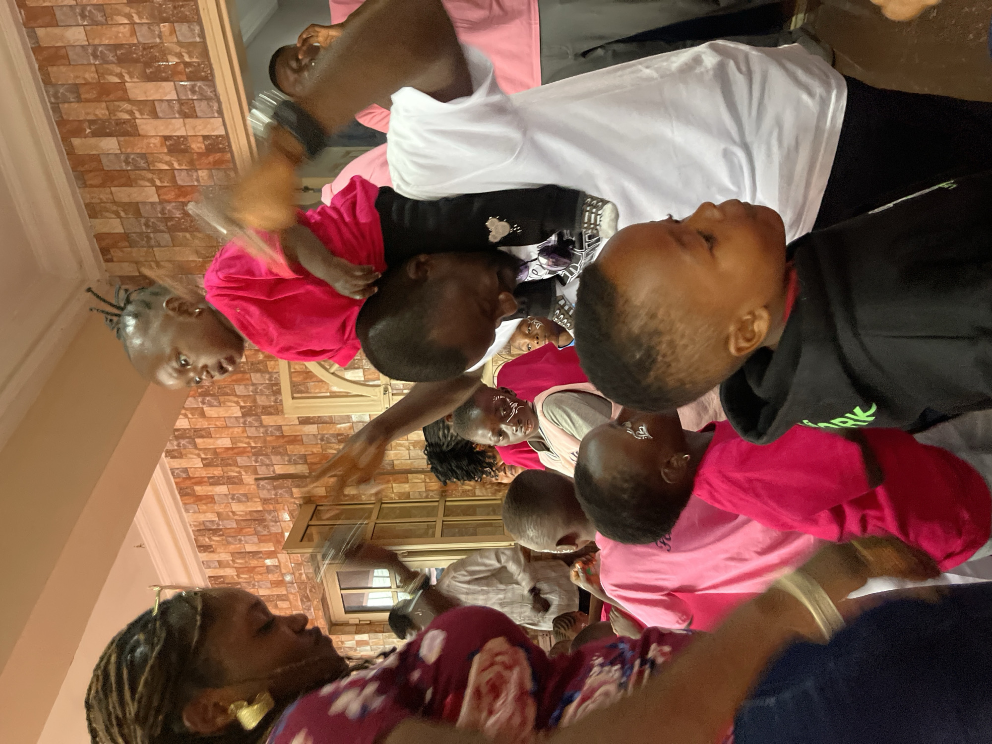 A group of people, primarily women and children, are gathered indoors, likely at a party or community event in Gura Top, Nigeria. The environment appears to be a decorated room with patterned wallpaper. A man is holding a baby on his shoulders, both wearing pink. Many children in the foreground have white face paint designs. Several individuals are wearing pink clothing, suggesting a themed event. The overall atmosphere seems festive and familial.