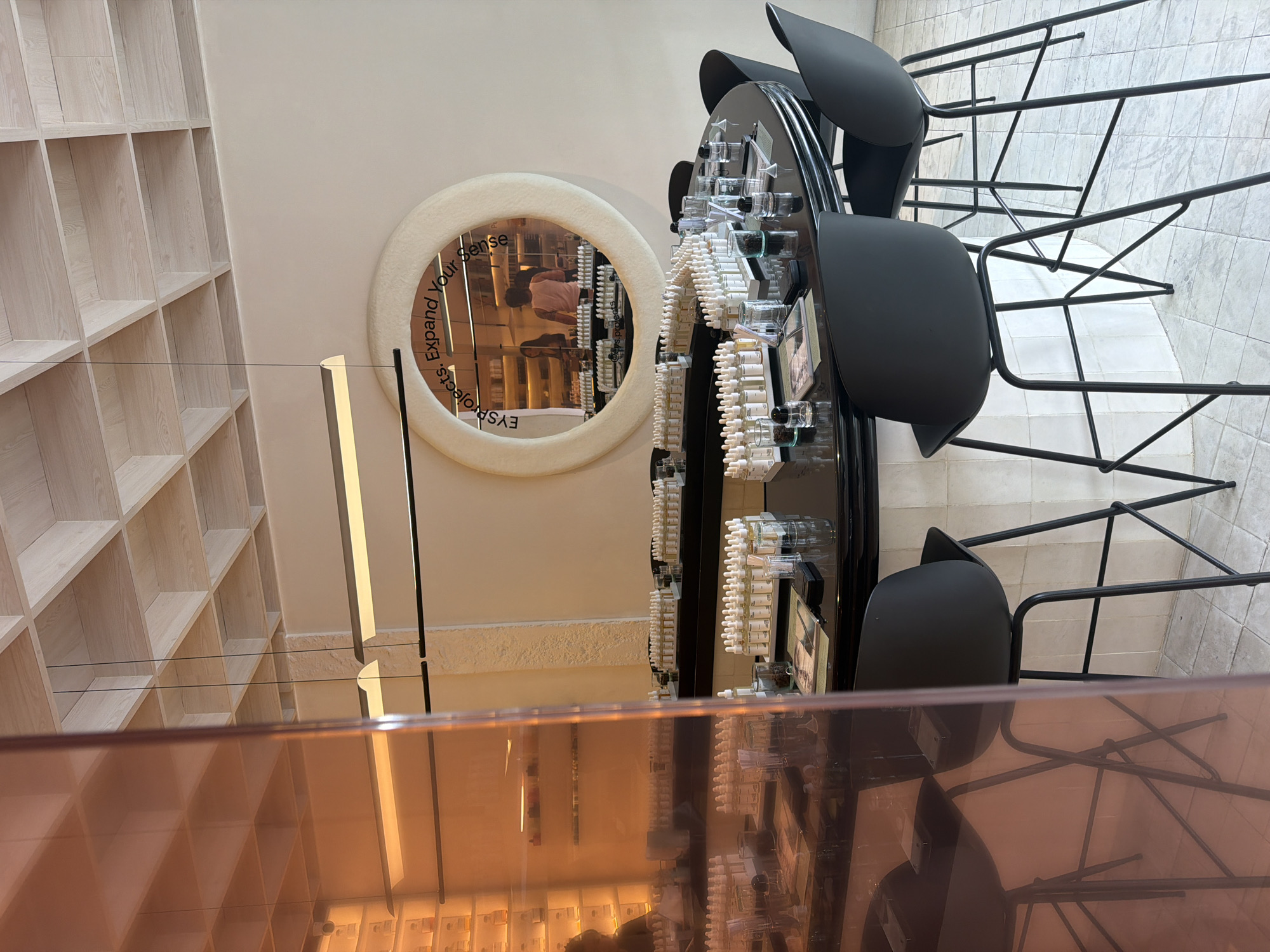 The image depicts the interior of a modern, upscale establishment, possibly a perfume boutique or a scent-testing laboratory, in Mengwi, Indonesia. The space features a circular counter with black stools, arranged around a central display of numerous small bottles, likely containing various scents. The counter itself is dark and reflective, showcasing more rows of these bottles, along with small glass containers and what appear to be samples.
Above the counter, a large, circular mirror with a textured, fuzzy frame is prominently displayed. The mirror reflects the interior, showing shelves lined with more bottles and two individuals standing in the background, one wearing a pink shirt and the other a dark top. Text is visible on the mirror's surface, reading "Evs projects. Expand Your Sense."
The ceiling is a distinctive architectural feature, composed of a grid of light-colored wooden squares, creating a contemporary and airy feel. Two minimalist pendant lights hang from the ceiling, casting a warm glow. The walls are a neutral, light color, and the floor is tiled with small, pale squares. To the left of the frame, a large, translucent orange panel partially obscures the view, reflecting the interior and adding a warm, amber hue to that section of the image. The overall ambiance suggests a sophisticated and inviting space focused on sensory exploration.