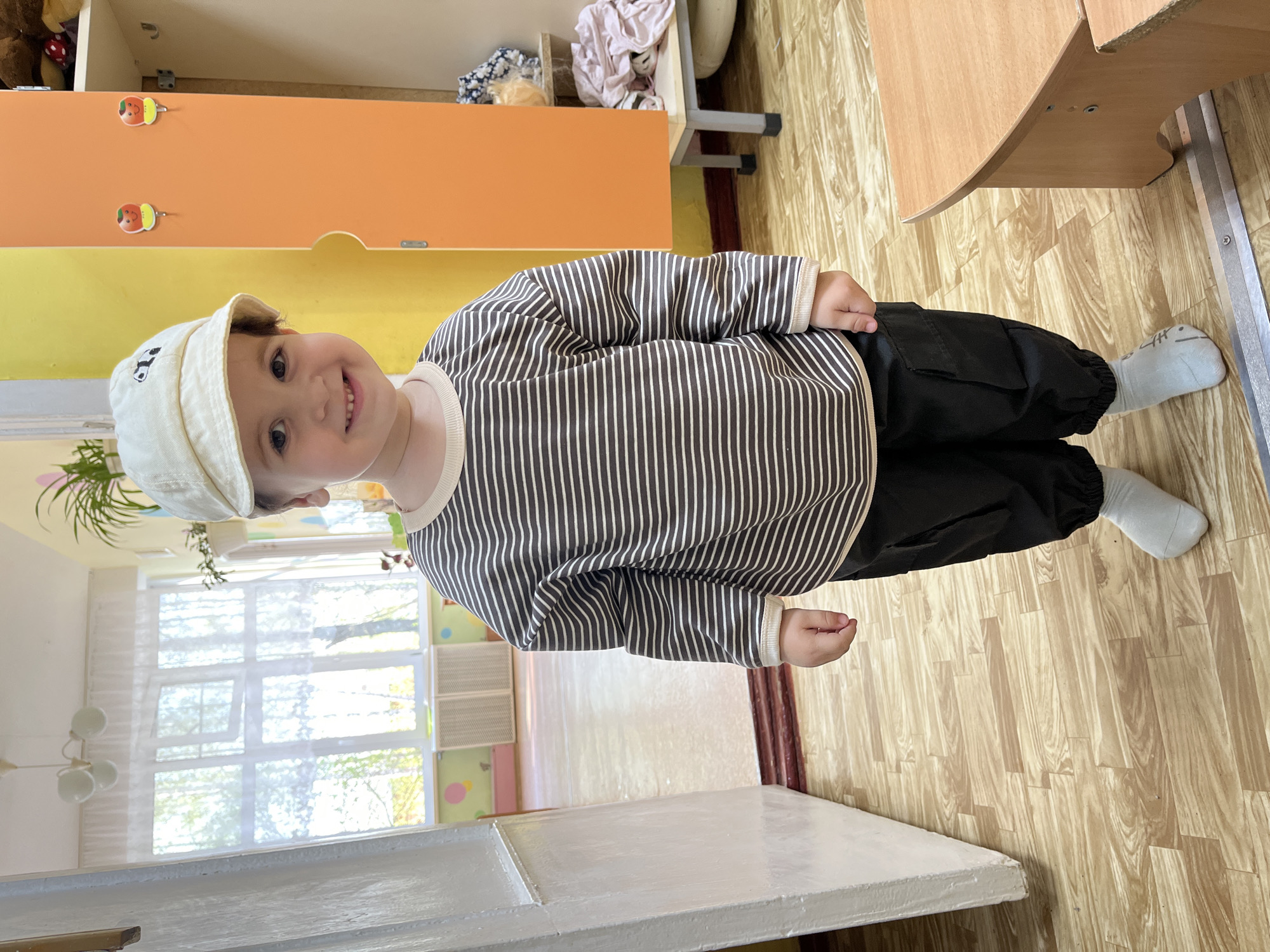 A child wearing a striped shirt, black pants, white socks, and a white cap stands indoors on a floor with a wood-like pattern. Behind the child is an orange and yellow cabinet with mushroom decorations on the drawers. A desk is partially visible to the right. In the background, a window lets in natural light. Based on image metadata, this photo was taken in Chaikovsky, Russia.