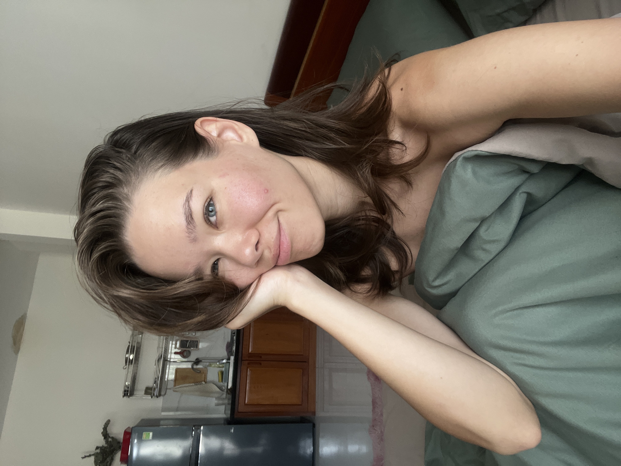 A young woman lies in bed under a green blanket. She rests her head on her hand, with her brown hair falling around her face. A wooden headboard is behind her.
In the background, a small kitchen area is visible, featuring wooden cabinets, metal shelving, a countertop with various items, and a black refrigerator. The room appears to be in an apartment or home in the city of Nam Nha Trang Ward, Vietnam.
