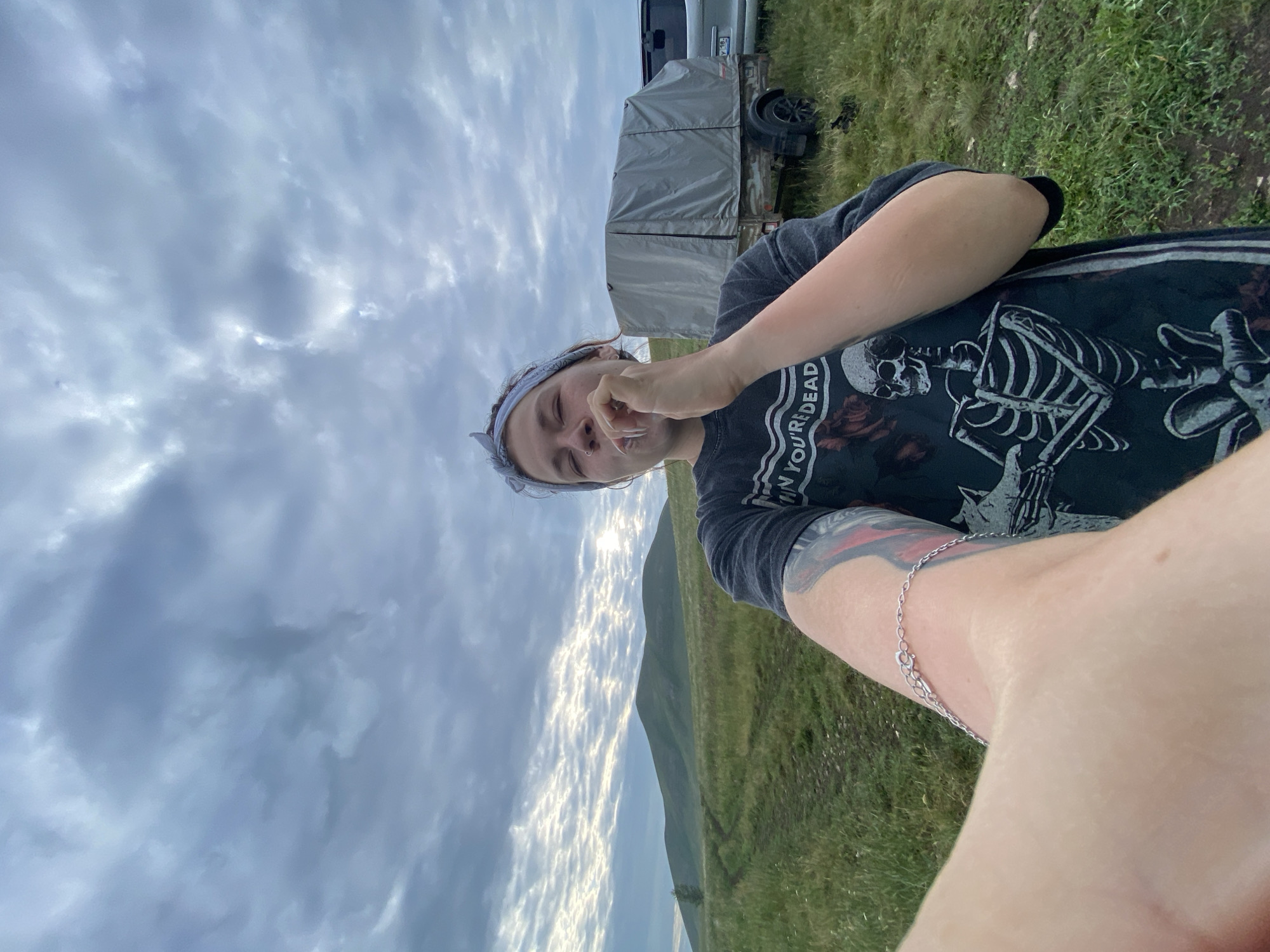 In a grassy field under a cloudy sky, a young woman is taking a selfie. She is wearing a dark t-shirt with a skeleton graphic and the text "WHEN YOU'RE DEAD". A light-colored bandana is tied around her head. Her right hand is raised to her mouth, holding a white object, possibly a cigarette or vape pen. A silver bracelet is visible on her left wrist, which is extended towards the camera. In the background, a gray-covered trailer and a portion of a silver vehicle are parked on the grass. Rolling hills are visible in the distance, with a single tree on the left side of the frame. The location is identified as the village of Абзаново, Russia.