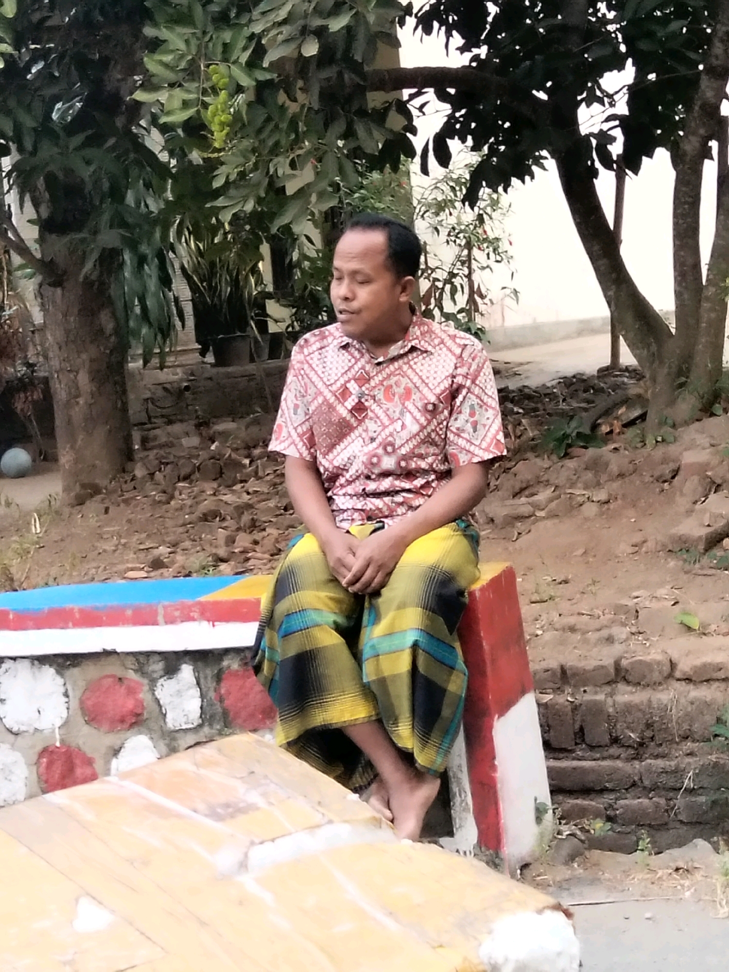In Jenangan, Indonesia, a man sits outdoors. He wears a patterned short-sleeved shirt and a sarong with yellow, blue, and black plaid. The man is seated on a red and white post. In front of him is a wooden surface, and behind him is a wall with red and white circular decorations. The background consists of trees, foliage, and a brick wall. The ground is covered in dirt and debris.