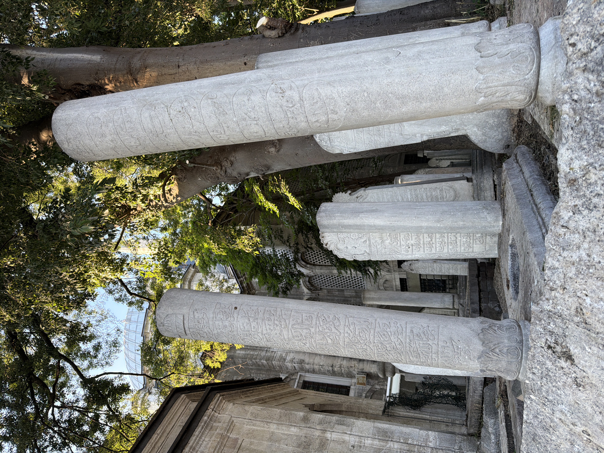 This image shows a graveyard with several tall, stone tombstones, some of which are inscribed with Arabic calligraphy. The tombstones are of various sizes and shapes, with some being cylindrical and others flat. The graveyard is surrounded by trees, and a building with a dome can be seen in the background. The setting appears to be the Eyüpsultan Cemetery in Eyüpsultan, Istanbul, Turkey, a historic and revered burial ground. The lighting suggests it is daytime, possibly late afternoon due to the long shadows. There are no people visible in the image, but the presence of tombstones indicates a place of burial. The environment is one of stillness and historical significance.