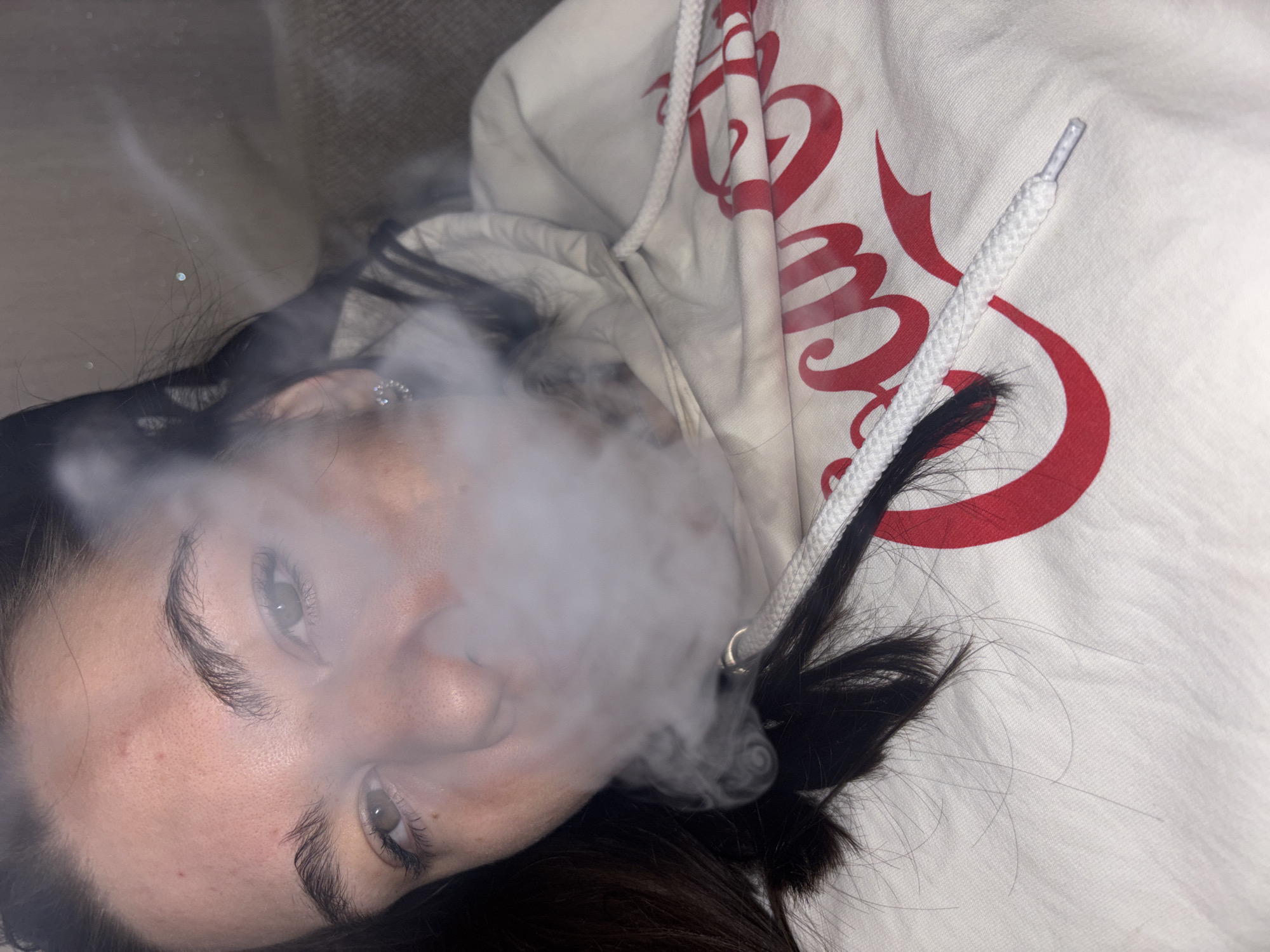 A woman with dark hair is lying down wearing a light-colored hooded sweatshirt with red lettering. She is exhaling a cloud of vapor or smoke. She has fair skin and visible eyebrows and eyelashes. An earring is visible in her left ear. The background is indistinct. There are no indicators that this image was taken in Perm, Russia.
