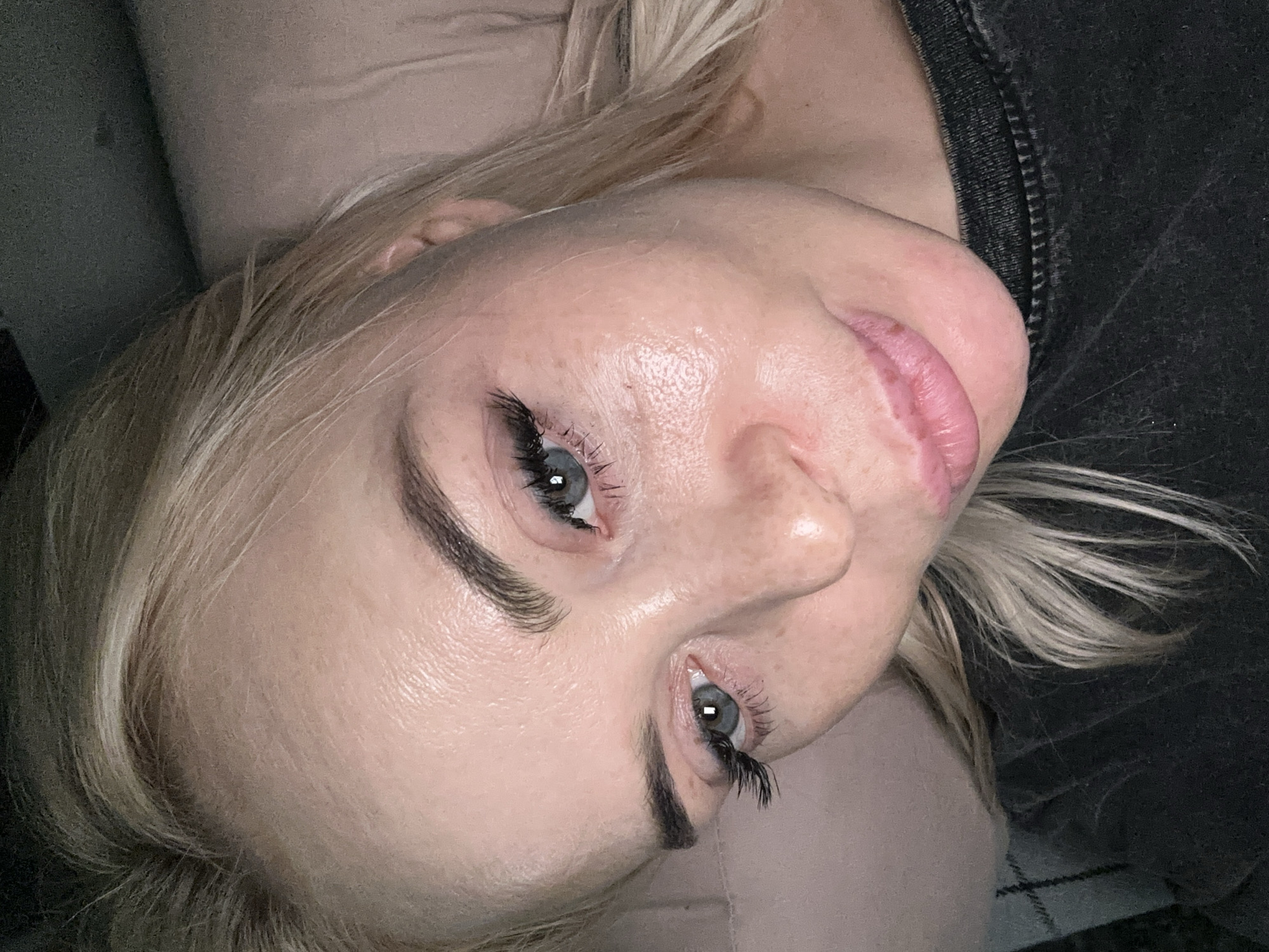 The media file features a close-up, high-angle shot of a woman with blonde hair, lying down and looking upwards towards the camera. Her head is tilted, giving the image a rotated perspective. She has fair skin, blue eyes, dark, well-defined eyebrows, and long, full eyelashes. Her lips are a natural pink shade, slightly parted, and appear somewhat chapped. Her blonde hair is casually styled, framing her face.
The woman is wearing a dark, possibly charcoal gray, top with a visible zipper detail on the right side of the frame. The background is indistinct and dark, suggesting an indoor setting with subdued lighting. Her skin has a slightly reflective or dewy appearance. No specific activity is captured beyond her looking directly at the camera. There is no visible text in the image. The overall impression is a personal portrait captured in soft, dim light.